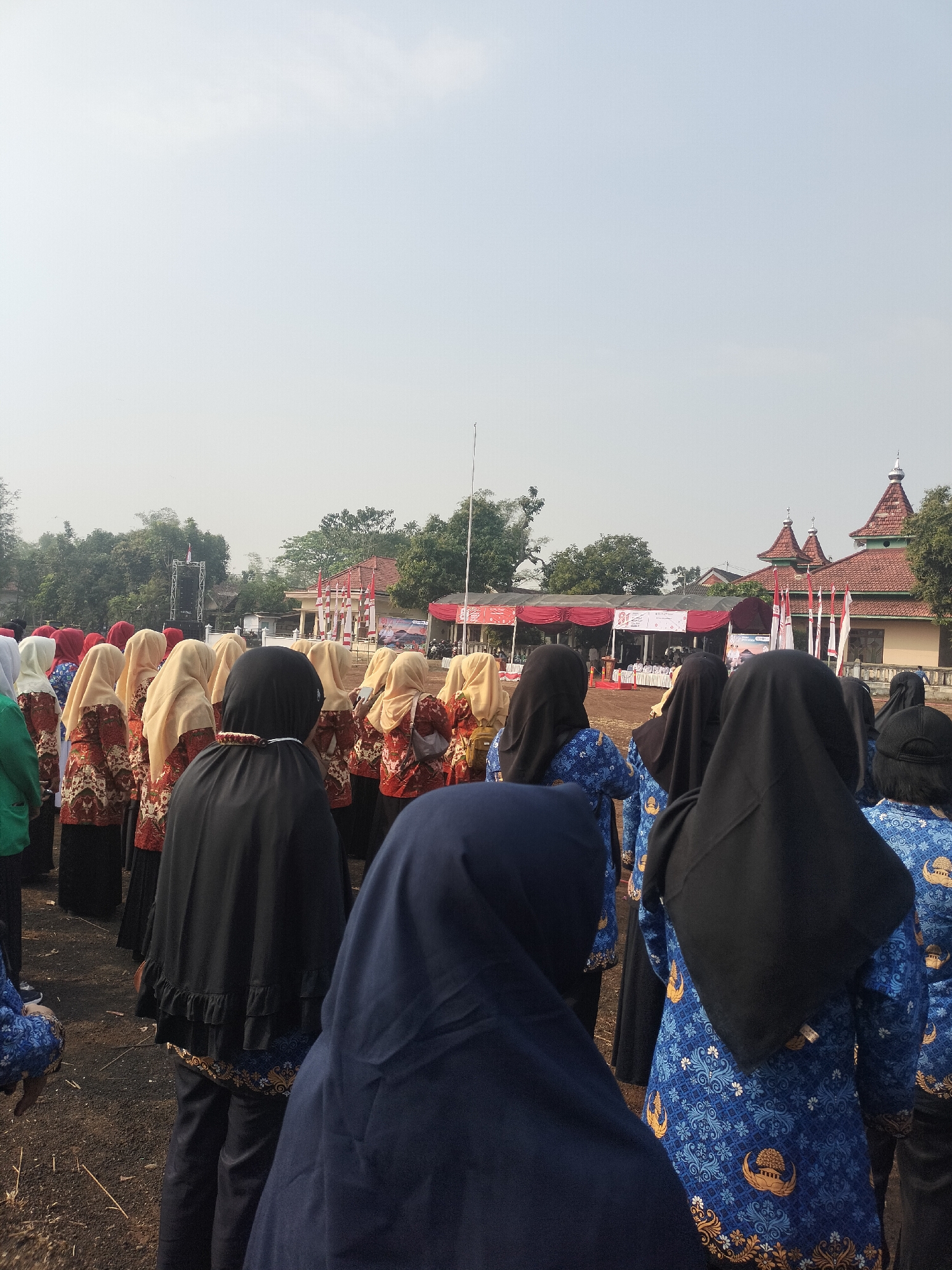 In Bantaran, Indonesia, a group of women in traditional attire are gathered outdoors. The women are standing in rows, facing away from the camera, with some wearing headscarves. Their clothing features a repeating pattern of gold crests and floral designs. The background shows a stage with red drapes, flags, and a large speaker system. Several buildings with tiled roofs and trees are visible beyond the stage. The ground is covered in dirt and small debris. The sky is overcast and bright.