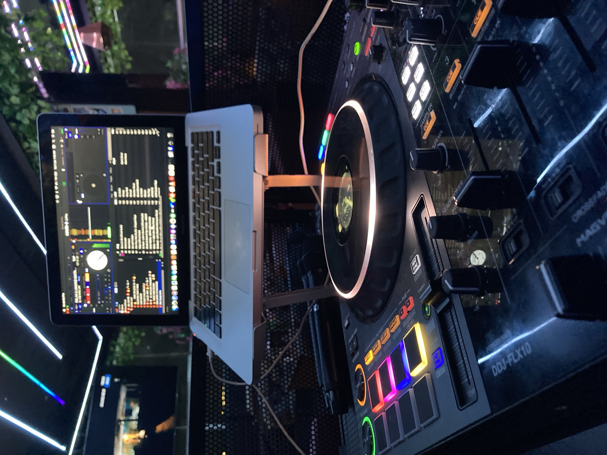 A DJ setup is shown in a dimly lit interior, possibly a bar or club, in Abuja, Nigeria. The scene focuses on a DJ controller with illuminated pads and faders, and a laptop displaying DJ software. The laptop is elevated on a stand. The background features colorful LED strip lights and some greenery, suggesting an outdoor or semi-outdoor setting. No people are visible in the frame.