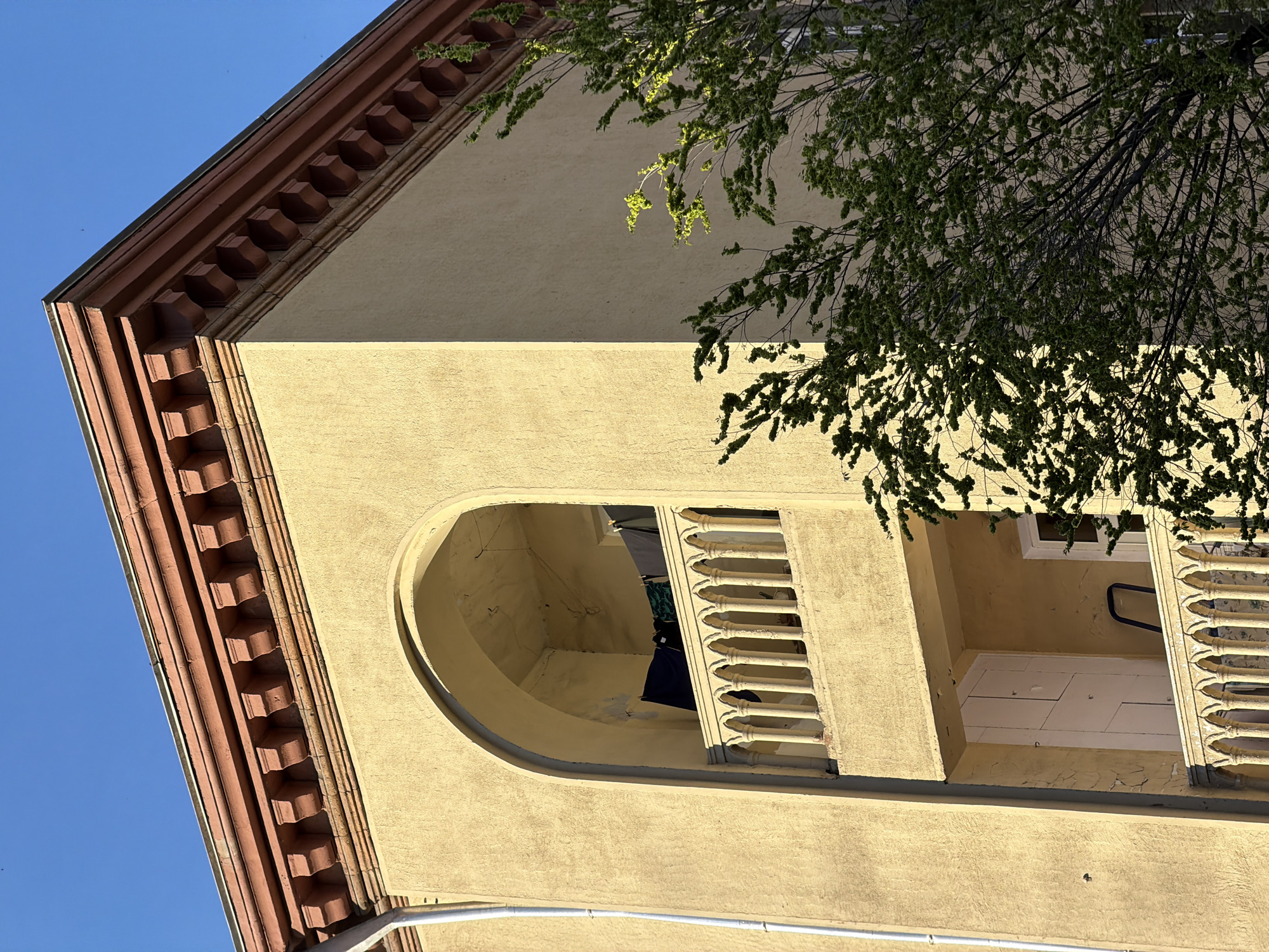 An upward-angled shot captures the corner of a building against a clear blue sky, partially obscured by the foliage of a tree. The building features a warm, light yellow or beige stucco facade with a prominent reddish-brown cornice and eaves. The cornice is adorned with a decorative row of dentil-like brackets. Below the main cornice, a deeply recessed, arched balcony or loggia is visible, featuring a classical balustrade with multiple light-colored balusters.
Inside the arched recess, signs of daily life are present, with dark blue and lighter fabrics hanging, possibly to dry. Parts of a white door or window and a black handle or railing are also discernible within the balcony area. The building's plaster shows some signs of age, with minor flaking visible. A white pipe or conduit runs horizontally across the lower section of the facade.
Direct sunlight illuminates the building, creating strong contrasts between light and shadow and highlighting the textures of the stucco and the architectural details. This suggests a clear, sunny day, likely in the morning or late afternoon due to the angle of the light. No people are visible. The scene depicts a quiet architectural detail of a building in Yerevan, Armenia. There is no visible text.