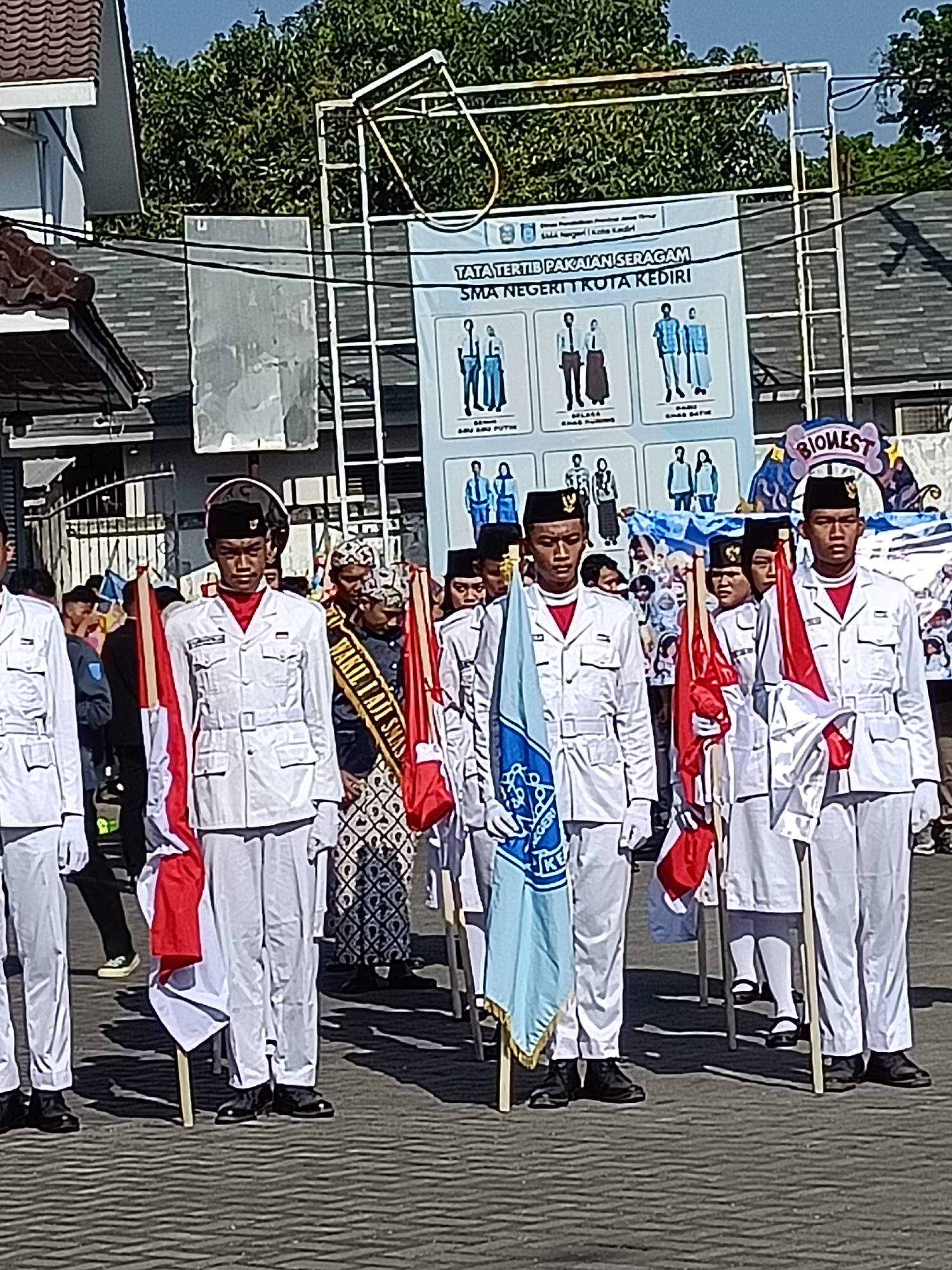 This image depicts a group of young people in uniform, likely a school or youth organization, participating in an event in Kediri City, Indonesia. The central figures are young men in white uniforms, black hats, and red scarves, carrying flags. One flag is light blue with white lettering and symbols. Another flag is red and white. They appear to be marching or standing in formation on a paved surface, possibly a street or public square.
Behind them, a large banner is visible with text in Indonesian, including "TATA TERTIB PAKAIAN SERAGAM SMA NEGERI KOTA KEDIRI," which translates to "Dress Code for Uniforms of State High School Kediri City." The banner also features illustrations of various uniforms. There are other individuals in the background, some wearing traditional attire, suggesting a cultural element to the event. The environment appears to be outdoors, with trees and buildings visible in the background, under a clear, sunny sky. The overall scene suggests a formal or ceremonial gathering related to education and potentially local culture in Kediri City.
