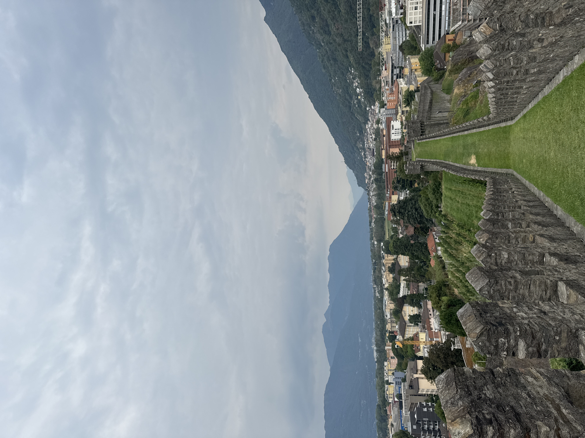 This is a vertical, eye-level, wide shot of the Castel Grande in Bellinzona, Switzerland, looking down a grassy path between two stone walls. The walls are topped with crenellations and appear to be made of rough-hewn grey stone. The path is a vibrant green, likely freshly mowed grass, and slopes gently downwards towards a city in the middle ground.
The city is a mix of modern and older buildings, with various colors and heights. Some buildings appear to be residential, while others might be commercial or industrial. Trees and greenery are interspersed throughout the city, and on the hillsides behind it. In the far background, large, dark, hazy mountains dominate the horizon, with a cloudy, overcast sky above them. The overall lighting suggests an overcast day, with diffused light and muted colors. There are no people visible in the image.