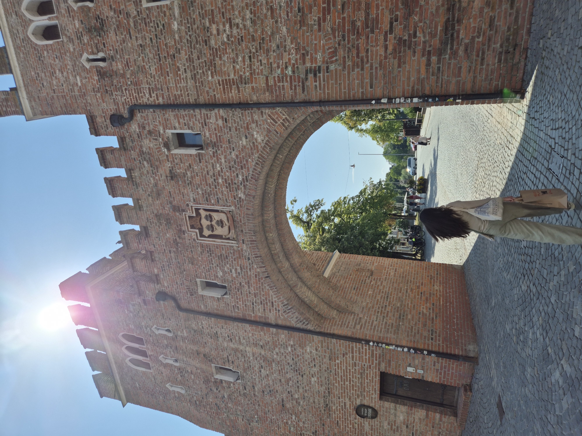 This image captures the Isartor gate in Landshut, Germany, on a sunny day. The ancient brick structure features a large arched entryway, crenellated battlements, and arched windows. A woman with long dark hair, dressed in a light-colored outfit and carrying two shopping bags, walks away from the viewer towards the arched opening. Beyond the gate, a cobblestone street is visible, lined with trees and with a few people and vehicles in the distance. Sunlight streams brightly from the upper left, creating lens flare and casting shadows on the cobblestones. A coat of arms is affixed to the outer facade of the gate above the archway.