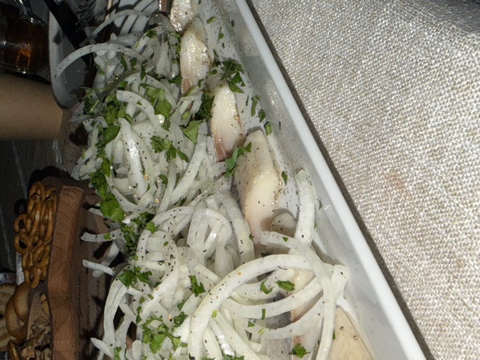 The image displays a close-up view of a dish featuring several pieces of what appears to be pickled or marinated fish fillets, generously topped with a large quantity of thinly sliced white onions. The entire dish is garnished with chopped green herbs, likely parsley or dill, and sprinkled with black pepper. The fish fillets are light in color, with a firm texture, and are partially visible beneath and alongside the translucent onion rings.
The dish is presented in a white, rectangular serving container or platter, which is positioned diagonally across the frame. To the right of the dish, a light-colored, textured fabric, possibly a linen tablecloth or placemat, is visible, adding a simple background texture. On the left side of the image, a dark wooden serving board is partially visible, holding other food items; specifically, some golden-brown, twisted snack items resembling small pretzels or baked goods are noticeable on the board. In the upper left corner, a portion of a dark glass bottle or container, containing a dark liquid, can be seen.
The lighting in the image appears somewhat subdued or indoor, casting some soft shadows. The photograph is taken from a slightly elevated, angled perspective, focusing directly on the food presentation. No text is visible in the image. The overall scene suggests a mealtime setting, possibly a casual gathering or a home-cooked presentation of a traditional dish.