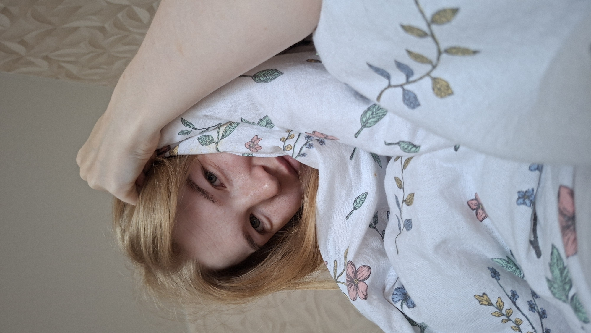 A close-up, upside-down shot captures a young person's face peeking out from under a white duvet with a floral pattern. The person has blonde hair and light-colored eyes, with a hint of a smile. Their left hand is visible, holding onto their hair or the duvet. The background appears to be a wall with a textured pattern. The lighting suggests it's daytime, and the scene is likely indoors, possibly in a bedroom, given the duvet. The overall mood is relaxed and intimate. There is no visible text in the image. The location context provided is Chaikovsky, Russia.