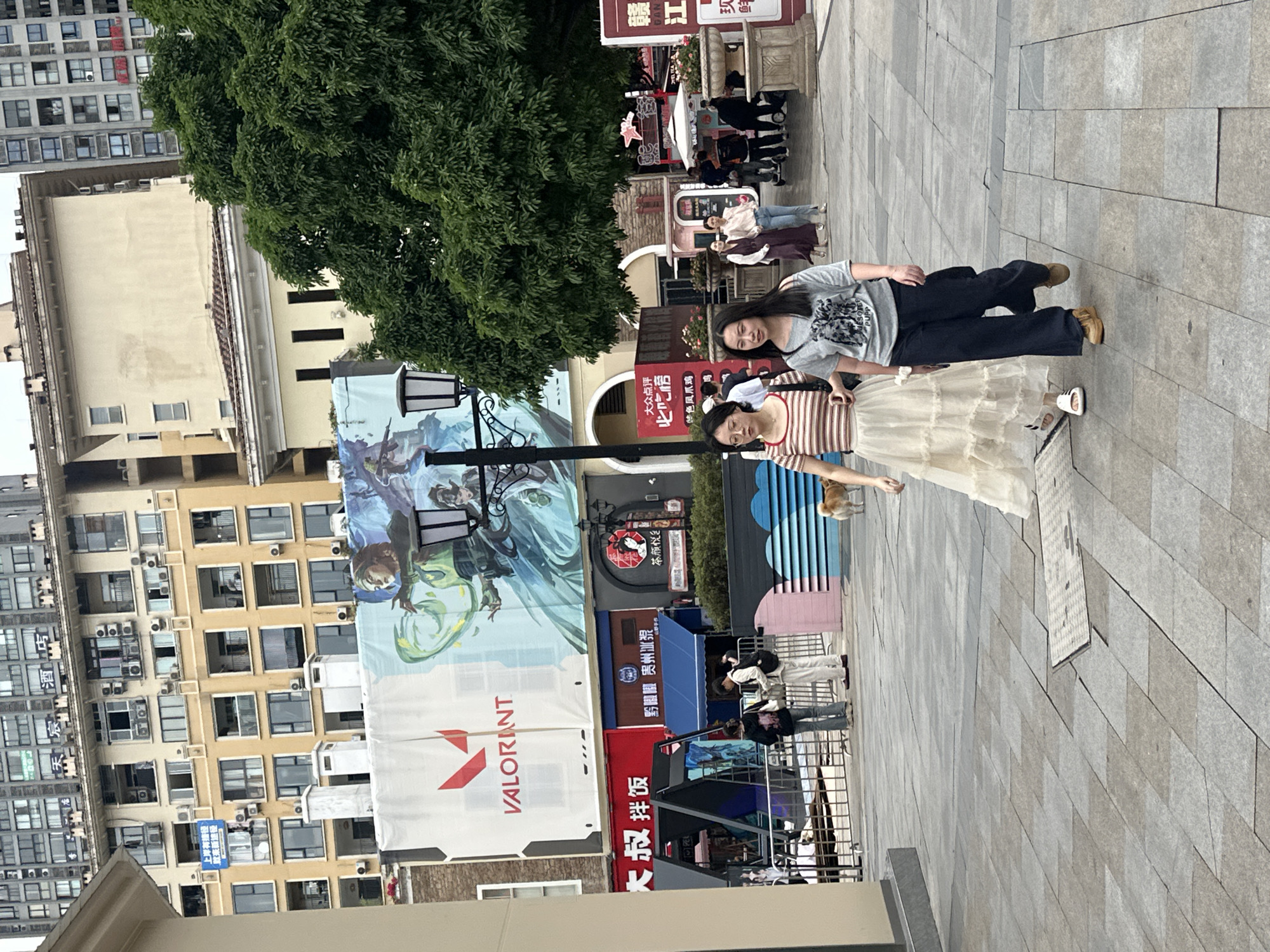 The image shows a public square, possibly in Chongqing High-tech Zone, China, given the Chinese characters visible. Two women stand prominently in the foreground. One woman wears a striped shirt and a light-colored, multi-layered skirt, with glasses perched on her face. The second woman is behind her, wearing a gray t-shirt with an image on it and dark pants.
Behind the women is a street with shops and advertisements. Prominent is a large banner advertising the video game Valorant. Other signs written in Chinese are partially visible. There is a large tree providing shade, and several buildings are visible in the background, including a multi-story apartment complex.
The ground is paved with rectangular stones. Some people are walking in the background. The lighting suggests it is daytime.