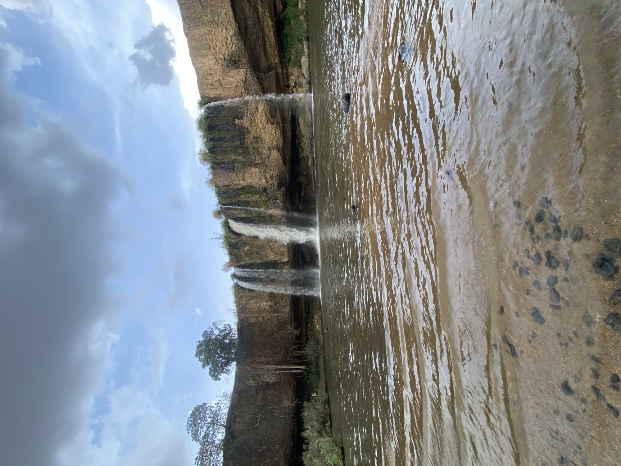 The image shows a waterfall cascading into a river or pool in Kukum, Nigeria. The waterfall is multi-tiered, with water flowing over rocky cliffs covered in some vegetation. The water appears murky. Two people are visible in the water. The right side of the image shows the riverbank with some scattered rocks. The sky is cloudy with patches of blue.