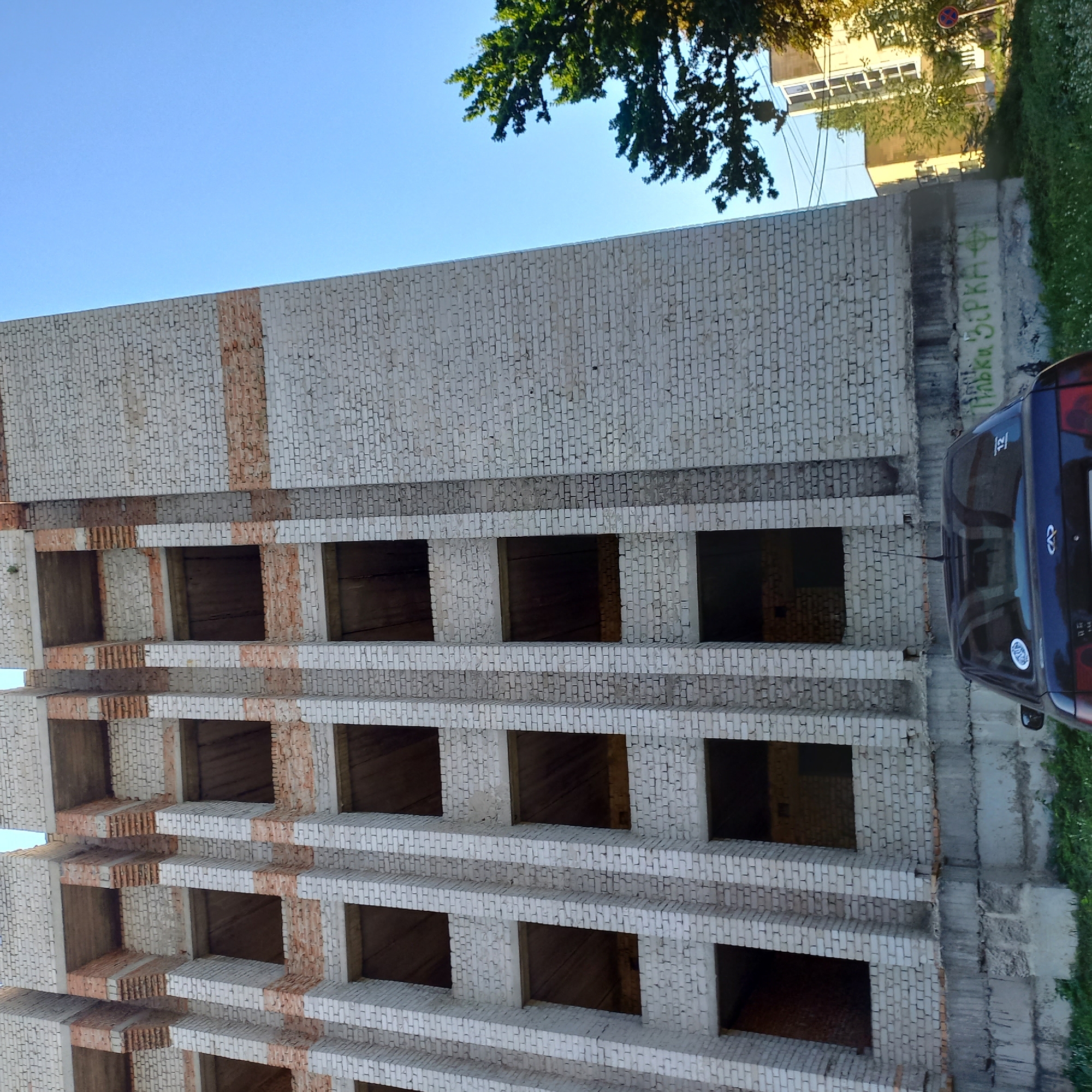 An unfinished brick building stands tall against a clear blue sky. The building's facade features a repeating pattern of white brick pillars interspersed with darker, recessed openings that appear to be windows or balconies. Some sections of the building reveal exposed concrete and brickwork, indicating construction is ongoing. In the foreground, a dark-colored sedan is parked. The car has a sticker on its rear window. The base of the building is concrete, with some green graffiti visible on it. Trees and other buildings can be seen in the background, suggesting an urban environment. The scene is well-lit by sunlight, casting subtle shadows. This image was taken in Kropyvnytskyi, Ukraine.
