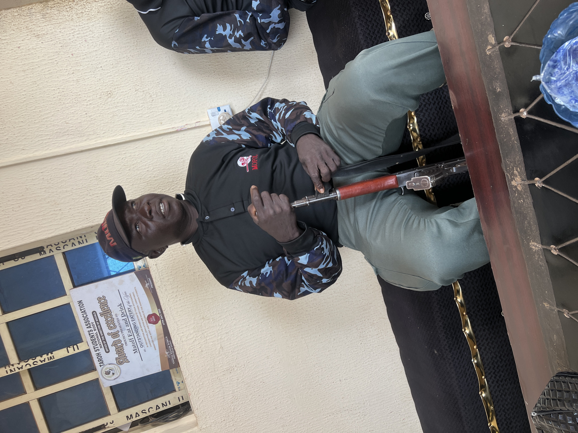 In Durumi, Nigeria, a man wearing a dark uniform with camouflage sleeves and a cap is seated. He is holding a rifle with a wooden stock, resting his hand on the trigger mechanism. The man has a dark complexion and appears to be looking towards the camera with a slight smile. In the background, a certificate is visible, awarding "Matoff Eat and Drink" the "Outstanding Eatery of the Year" award for their exceptional commitment and contribution. The certificate is dated July 27, 2025, and signed by the President, Nimnan Sansak Jemkur. The setting appears to be an outdoor or semi-outdoor area with a textured, light-colored wall and a dark, textured surface behind the man, possibly a bench or seating area. A wooden structure is also present to the right, with a blue plastic bag partially visible. The overall scene suggests an informal gathering or presentation.