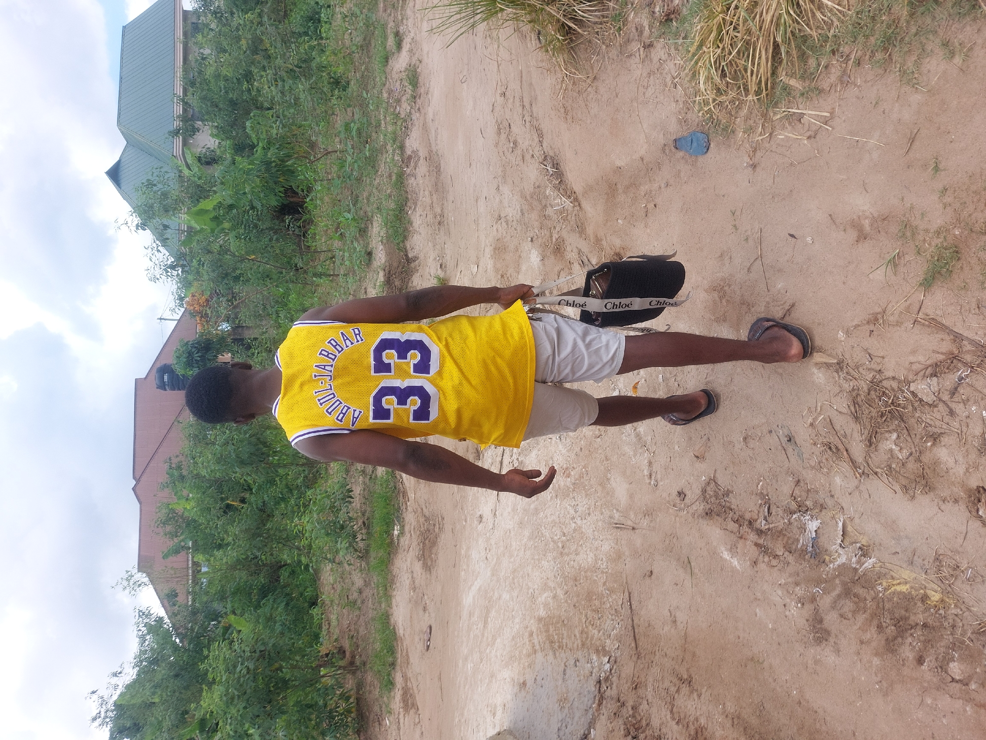A person wearing a yellow basketball jersey with the name "ABDUL-JABBAR" and the number "33" on the back is standing on a dirt path. They are wearing light-colored shorts and sandals. The person is holding a black bag with white straps in their left hand. The path is surrounded by greenery, and there are buildings visible in the background. The exact location within Dar es-Salaam, Tanzania, is not discernible from the image.