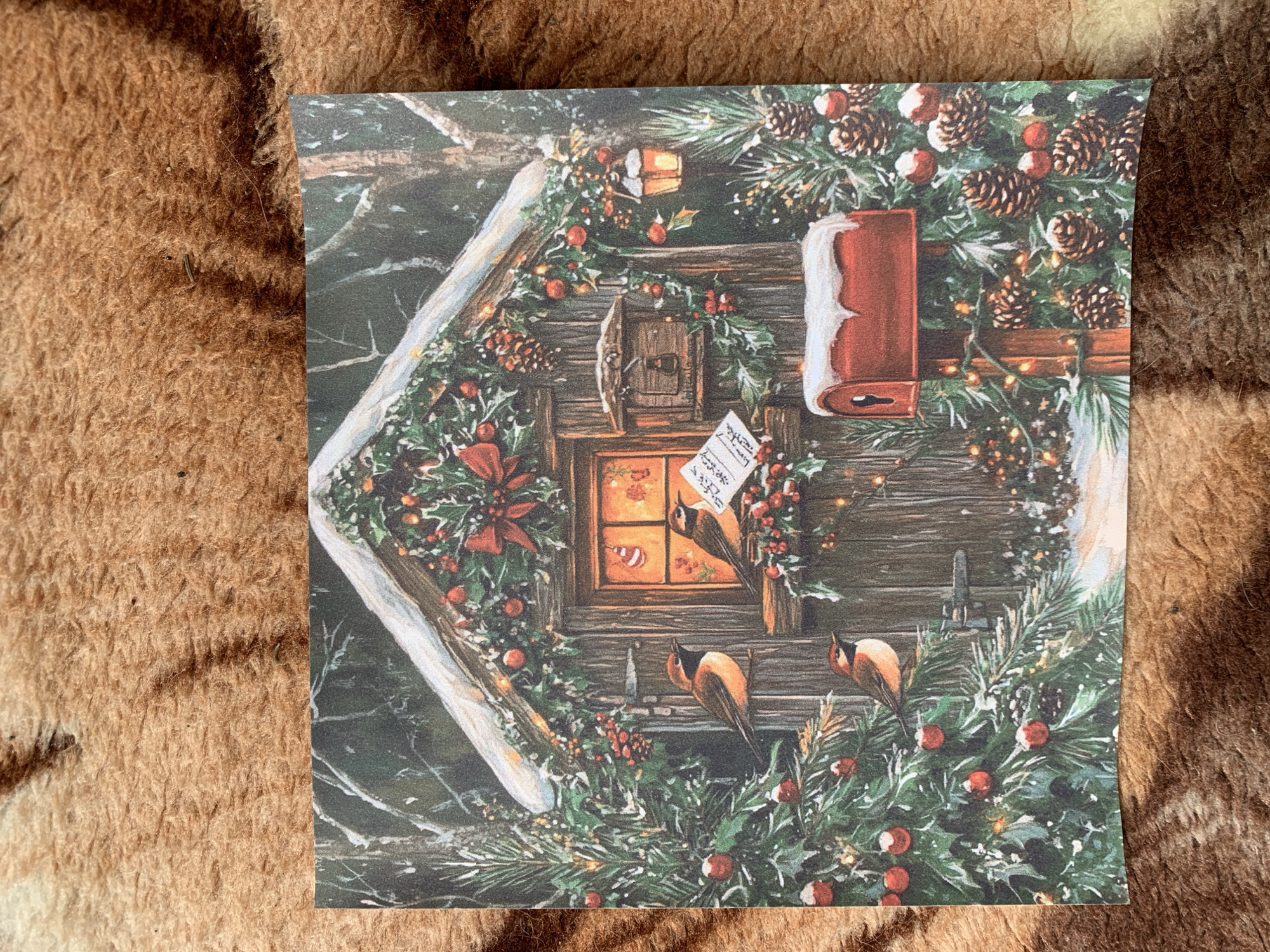 The image shows a square card depicting a winter scene, possibly a cabin or building decorated for Christmas. The building's facade is wooden and adorned with pine branches, pinecones, holly with red berries, and small lights. A red mailbox, partially covered in snow, is visible. A window glows warmly, and a note is hanging on the wall. Two birds perch on branches. The image background is a brown, furry surface.
The city of Slavyani, Bulgaria is not discernible in the image.