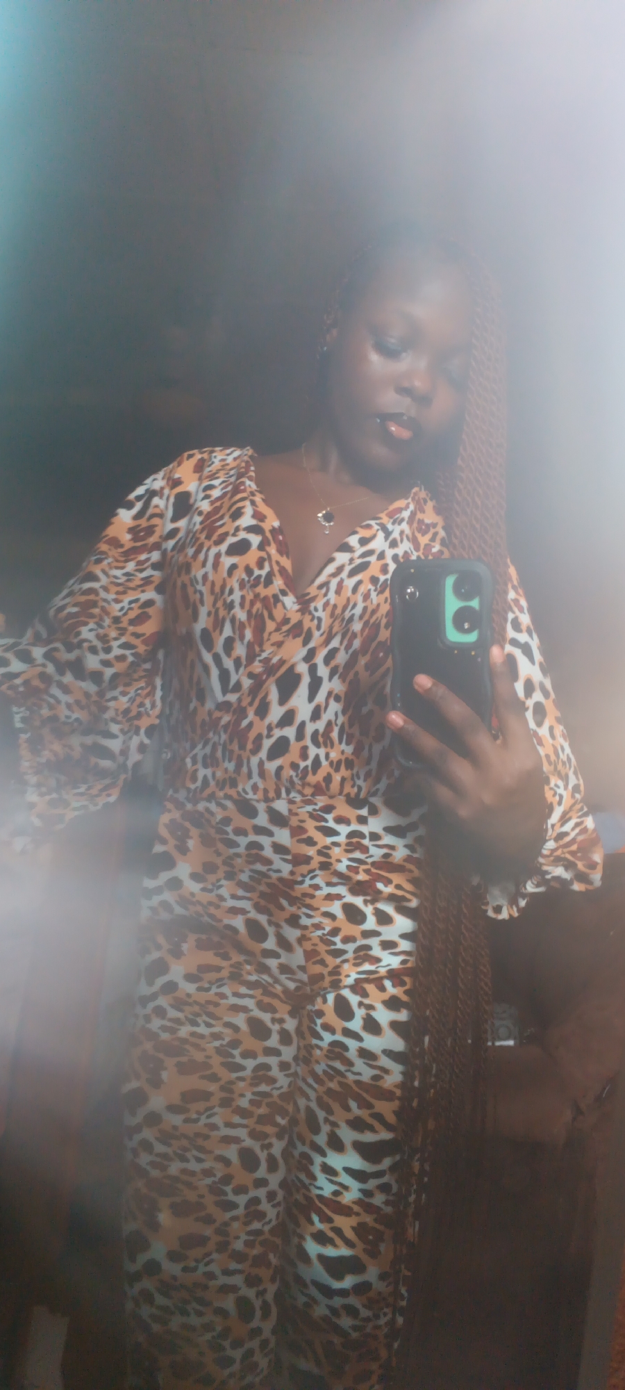 A young Black woman is taking a mirror selfie. She is wearing a leopard-print jumpsuit with flared sleeves and a V-neckline. She has long, braided hair and is wearing a necklace. She is holding a smartphone in her right hand, which is angled to capture her reflection. The background is dark and appears to be indoors, possibly a bedroom, with a brown armchair visible to the right. The lighting is dim, and there is a hazy, bright light emanating from the left side of the frame, creating a diffused effect over the image. The scene is consistent with a private residential setting in Onitsha, Nigeria.