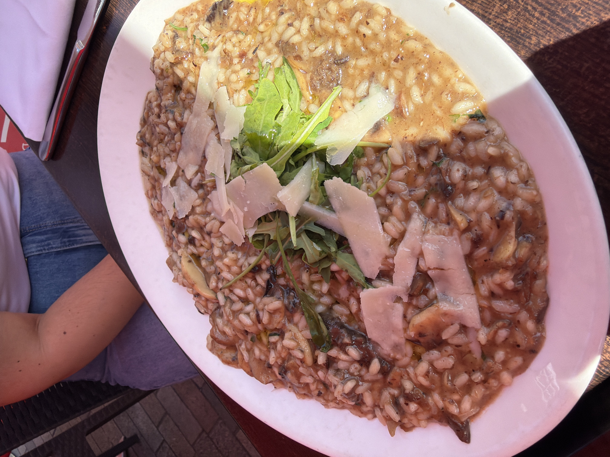 A high-angle, full-frame shot shows a plate of risotto on a dark, possibly wooden, surface. The risotto is garnished with arugula and shavings of parmesan cheese. A person's arm and leg are visible on the left side of the image, suggesting the photograph was taken by someone seated at the table. A napkin and a metal object, possibly cutlery, are also partially visible next to the arm. The location details (Guildford, UK) are not discernible from the image alone.