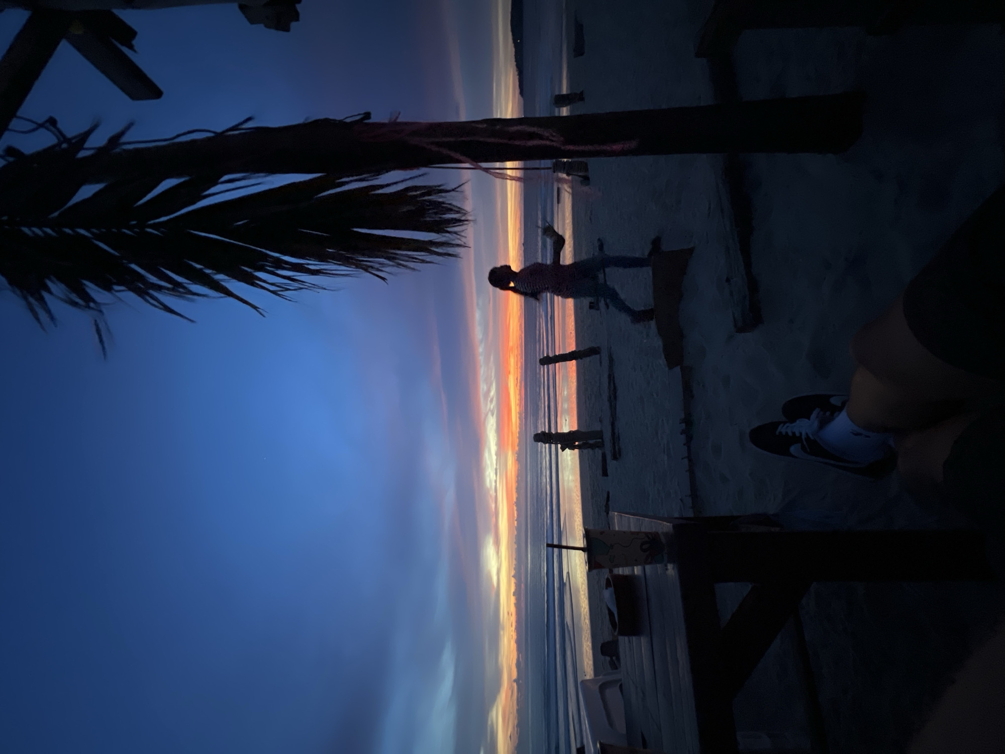 The image depicts a beach scene at dusk. The sky is a gradient of deep blue, transitioning to shades of orange and pink near the horizon, indicative of a sunset. Waves are visible on the ocean, reflecting the colors of the sky. In the foreground, a dark wooden table and chairs are present, with a person's legs and feet wearing sneakers and white socks resting on it. To the right, a silhouetted palm tree stands tall, its fronds reaching towards the sky. A person is walking on the sand in the middle ground, silhouetted against the bright sky. Several wooden posts are scattered on the beach, some appearing to be part of structures. The overall atmosphere is tranquil and serene.