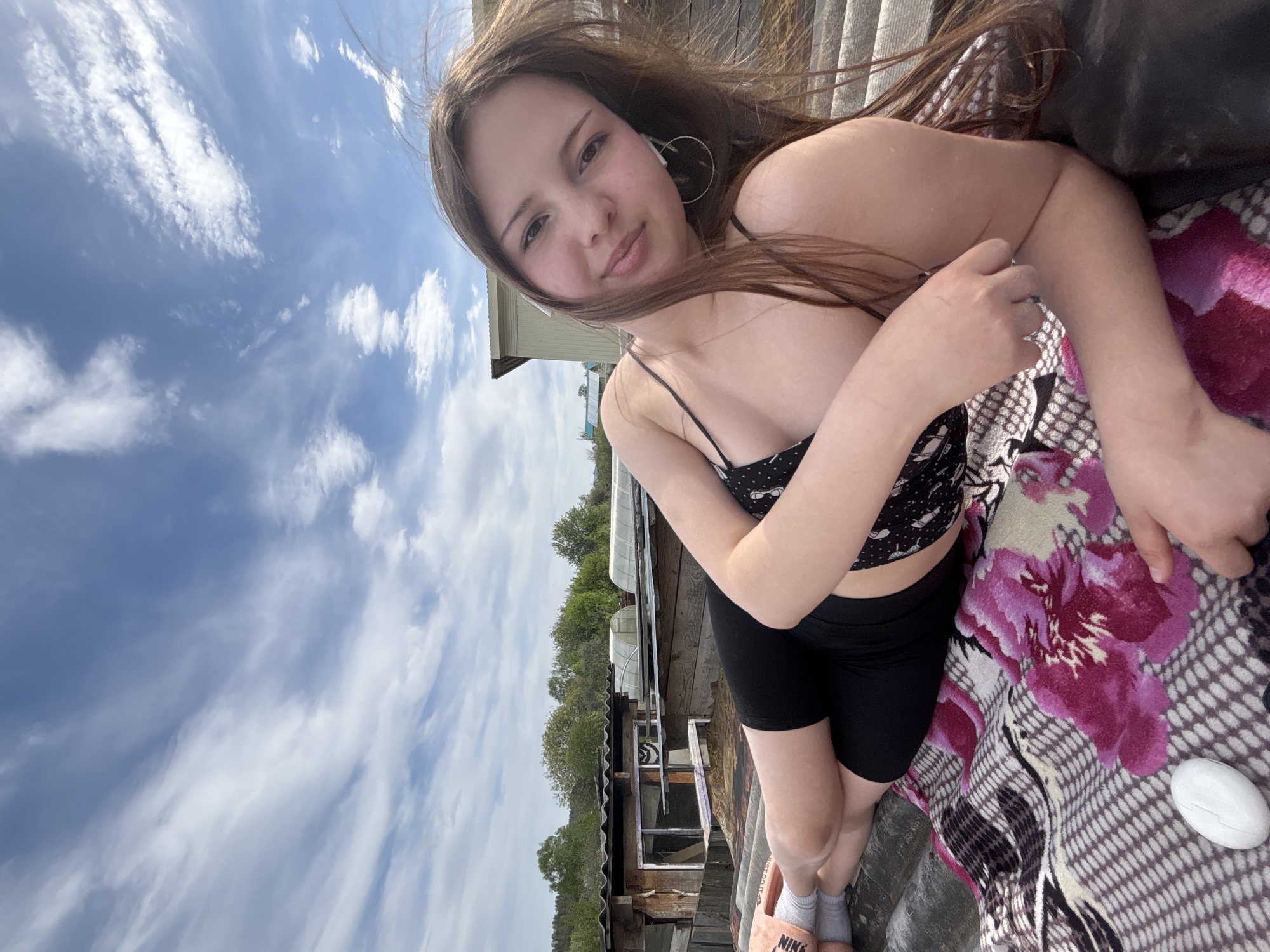 A young woman lies on a patterned blanket on a rooftop in Фоки, Russia, on a sunny day. She is wearing a black patterned crop top, black shorts, and peach-colored slides with gray socks. Her long brown hair is loose, and she is wearing hoop earrings. In the background, there are trees, buildings, and a blue sky with scattered clouds.
