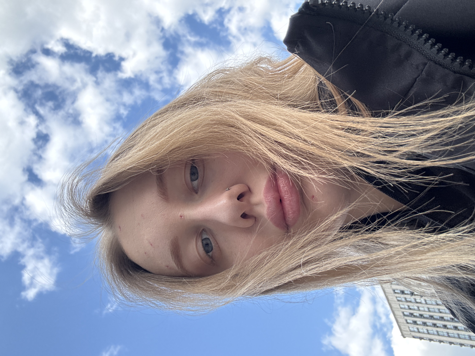 The image is a close-up, low-angle selfie, heavily rotated 90 degrees clockwise. It features a young person with long, flowing blonde hair, light-colored eyes (blue or grey), and full lips. The person has a neutral to slightly pensive expression, looking directly towards the camera. A small nose piercing is visible on her right nostril. She is wearing a black jacket, indicated by the visible fabric and zipper details around her shoulder and upper arm.
The background consists primarily of a clear, bright blue sky with scattered white, fluffy cumulus clouds, indicating a daytime setting with pleasant weather. In the bottom right corner (when the person is oriented upright), a portion of a modern, tall building with multiple windows is visible, suggesting an urban environment. The perspective is from below, looking up towards the subject and the sky. No text is visible in the image.