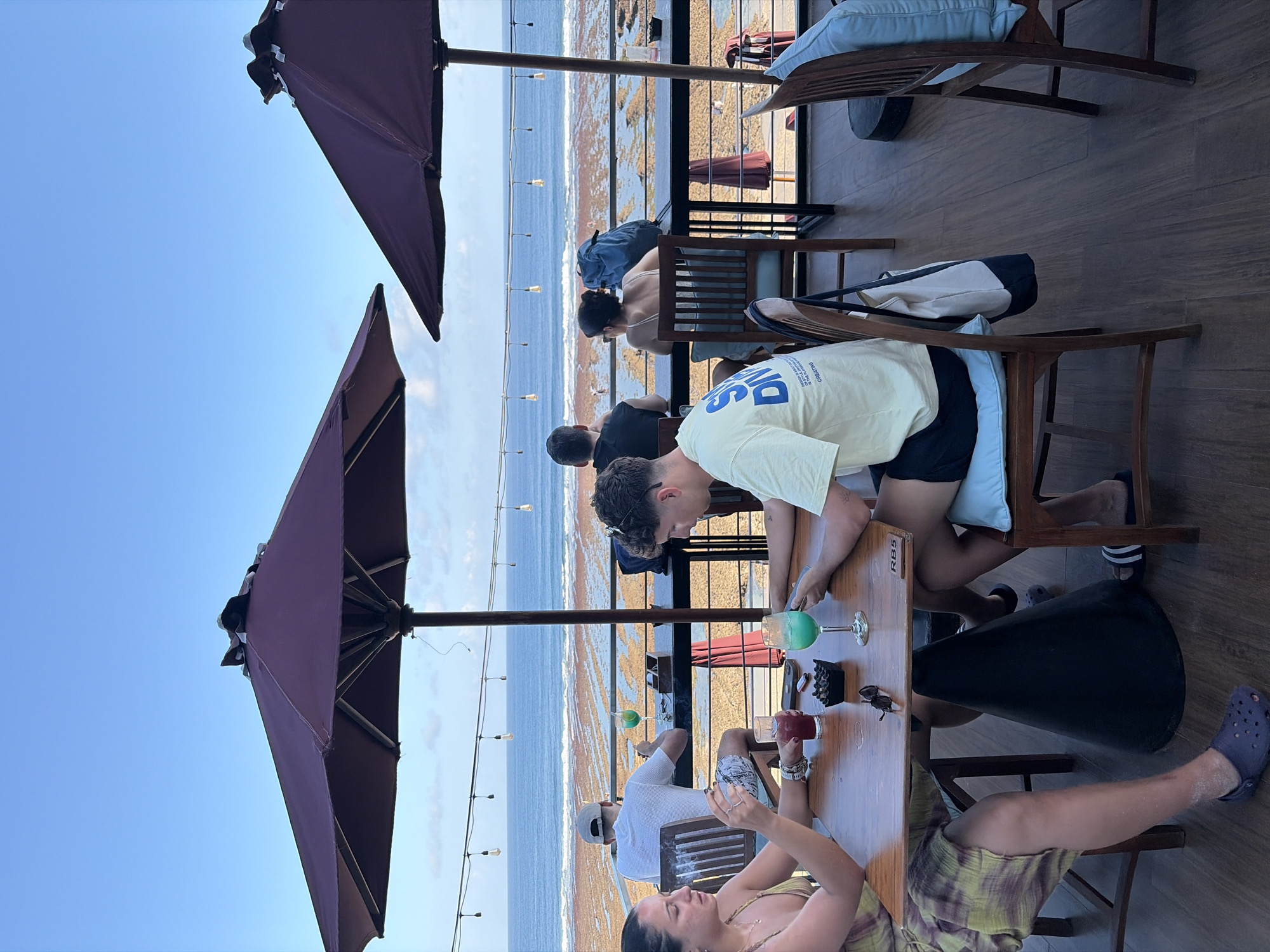 This image depicts a group of people seated at tables on an outdoor deck overlooking a rocky shoreline and the ocean. The location appears to be a restaurant or cafe in Kuta Selatan, Indonesia, given the style of architecture, the tropical ocean view, and the overall atmosphere.
In the foreground, two individuals are seated at a wooden table. One person, a woman, is wearing a patterned dress and has dark hair. She is holding what appears to be a cigarette and is looking towards her companion. The other person, a man, is wearing a light-colored t-shirt with graphics on the back and dark shorts. He is looking down at his phone. On the table between them are two drinks, one a vibrant blue, and an ashtray.
Behind them, a man in a white t-shirt and a cap is seated at a similar table, facing away from the camera, and appears to be looking out at the ocean. Further back, a woman with dark hair is seated with a blue backpack beside her. In the distance, the ocean stretches out under a clear blue sky with a few scattered clouds. String lights are hung across the deck. The deck itself is made of wooden planks, and there are several large, dark umbrellas providing shade. The rocky shoreline is visible in the mid-ground, with waves gently breaking. The overall impression is one of a relaxed, coastal dining experience.