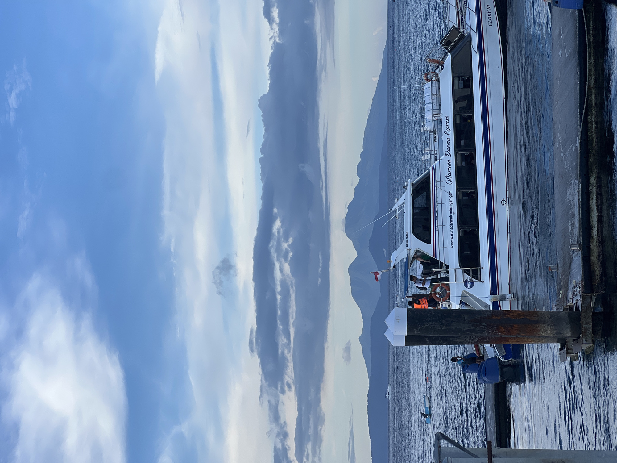 The image captures a serene waterfront scene at Kampung Toyapakeh, Indonesia, featuring a passenger boat docked beside a pier. In the foreground, sturdy wooden and metal pilings of a dock are visible, with calm, rippling water surrounding them.
The central subject is a white and blue striped fast boat, clearly marked with "Maruna Darma Express" on its side. Below this, the website "www.warunadarmalembongan.com" is displayed, hinting at its travel route, likely to Nusa Lembongan. Towards the bow of the vessel, the name "GLORY III" is also visible, along with a small red and white flag, possibly representing Indonesia. The boat features multiple dark-tinted windows for passengers and carries life rafts secured on its upper deck.
Several people are visible on and around the boat. Some passengers can be seen through the windows, while a person in an orange life vest is on the boat's deck near the dock. A smaller blue boat or dinghy is tied to the pier, with two individuals occupying it. Further out in the water, a lone person in a small kayak or similar craft is visible, along with other distant floating objects.
The background features a long range of hazy mountains stretching across the horizon, separating the vast expanse of water from the sky. The sky is partially cloudy, displaying patches of light blue interspersed with soft, elongated white-grey clouds, suggesting either early morning or late afternoon light. The overall scene conveys a calm, functional atmosphere associated with coastal transport and travel.