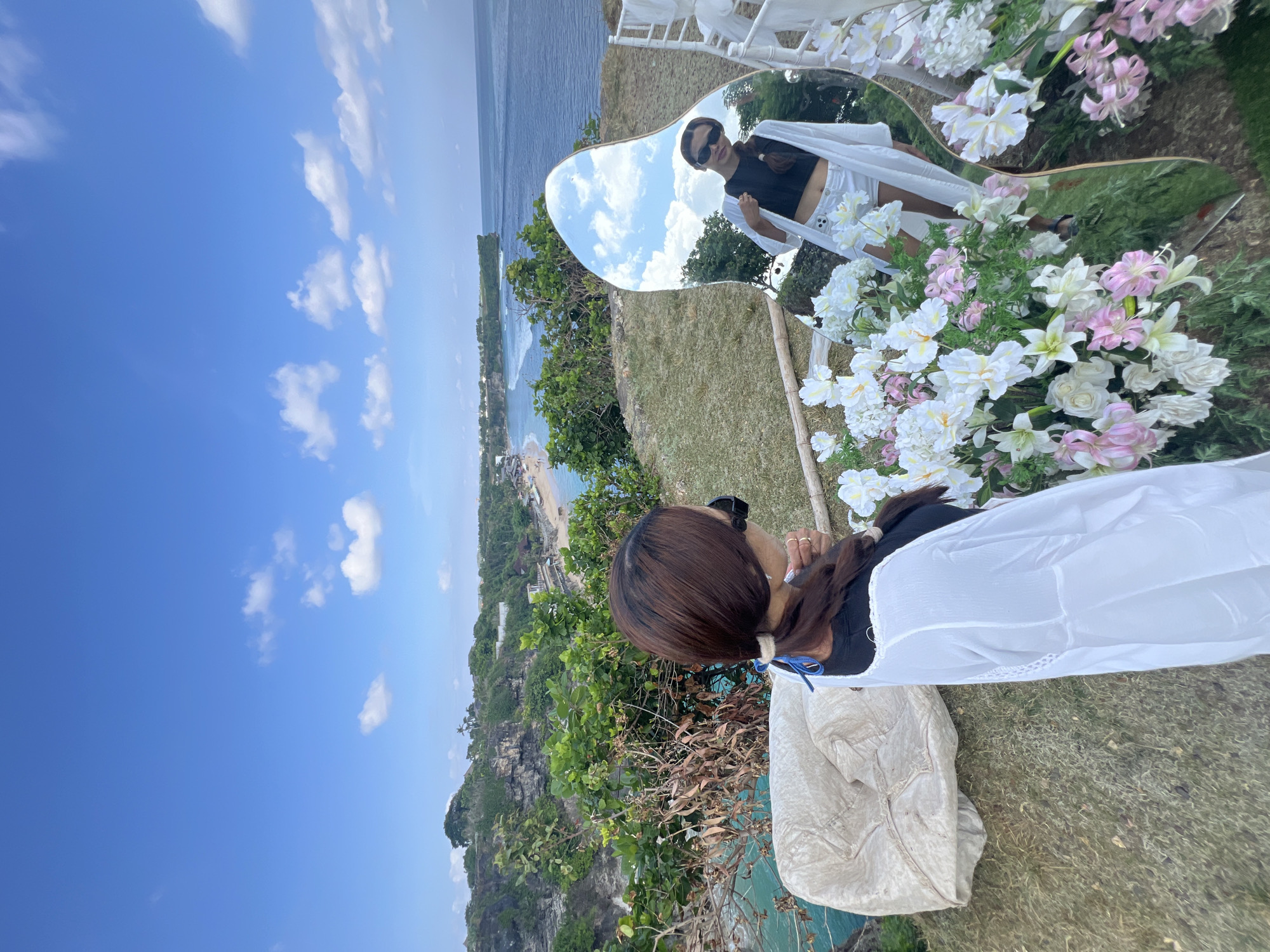 An outdoor image captures a scenic coastal view featuring a woman, a decorative mirror, and floral arrangements. The setting is a cliffside overlooking a vast turquoise ocean and distant sandy beaches, consistent with Kuta Selatan, Indonesia.
In the foreground, a woman with long, dark hair tied in a ponytail is seen from behind, wearing a black top and a sheer white cover-up. She appears to be gazing out at the expansive view. To her right, a prominent heart-shaped mirror stands adorned with an abundance of white and pink flowers, forming a decorative frame.
The mirror reflects the woman from the front, revealing her wearing sunglasses, a black crop top, and white shorts. It also clearly shows the bright blue sky with scattered white clouds. Partially visible white chairs are present near the mirror, suggesting a styled setup for an event or photoshoot. The ground is covered in green grass.
Beyond the immediate foreground, the landscape drops dramatically to the sea, characterized by lush green cliffs and vibrant turquoise water where waves crash onto sandy shores. The coastline extends into the distance under a clear, sunny sky, indicating daytime with excellent weather. There is no visible text in the image.