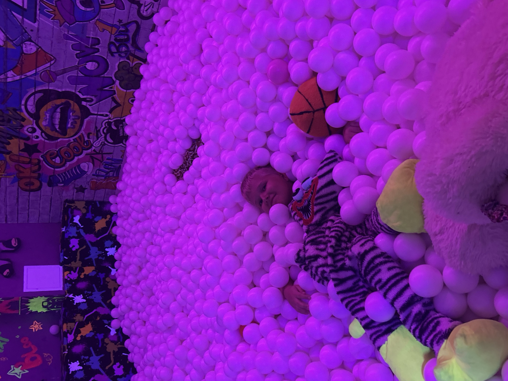 A young child is partially submerged in a ball pit filled with white balls under purple lighting. The child is wearing a black and white striped costume and is lying on their back. To the left of the child, a large stuffed animal with tiger stripes and yellow feet is visible. In the middle of the ball pit, an orange basketball is partially visible. The background features a wall with colorful graffiti art. The location is not specified as Chaikovsky, Russia, and this information cannot be inferred from the image.