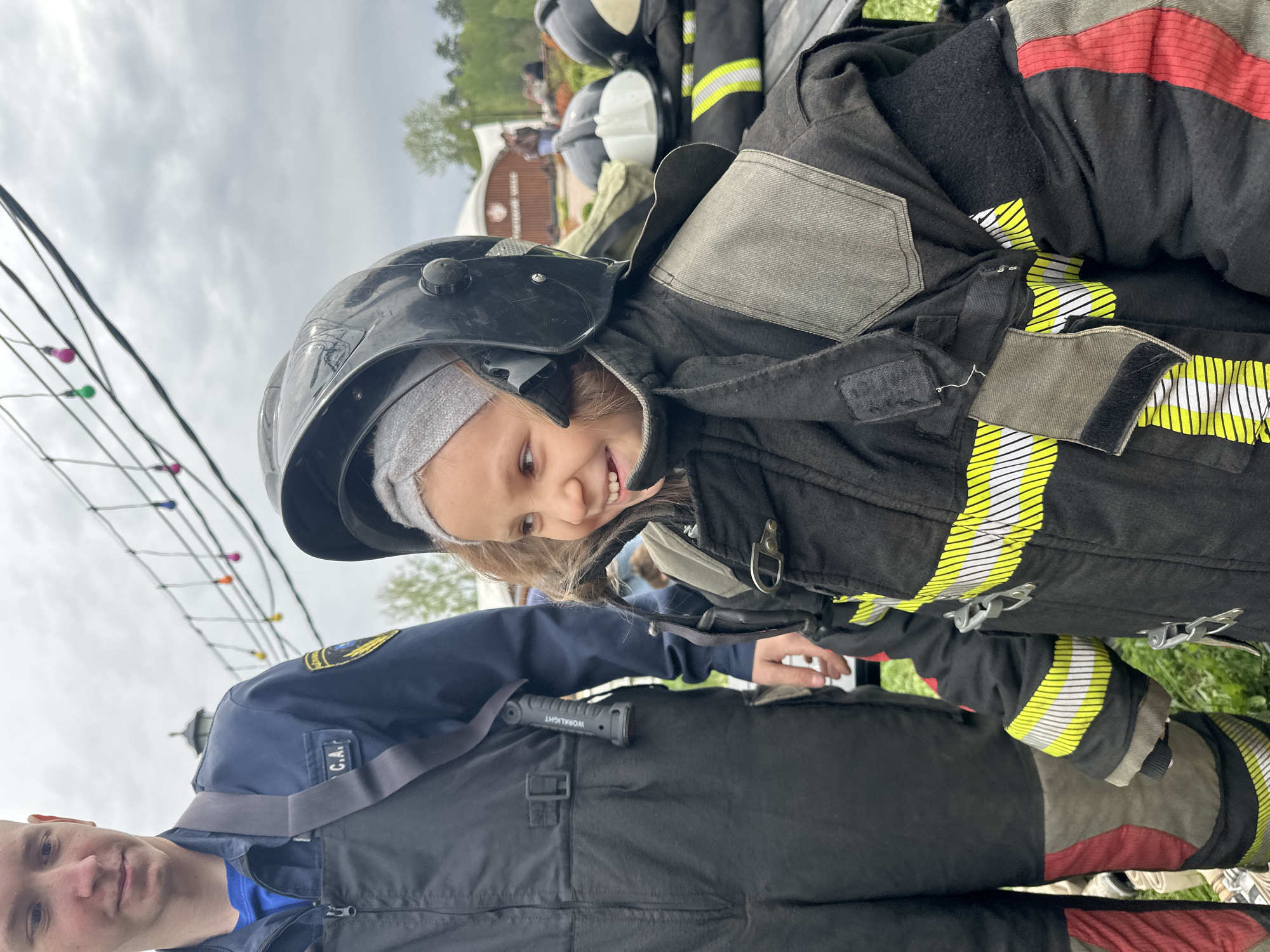 A young child wearing a black firefighter's helmet and a firefighter's coat poses with a man. The child is smiling and looking down and to the right. The firefighter's coat is black with yellow reflective stripes. The man in the photo is wearing a blue shirt and black overalls. "WORKLIGHT" is written on a cylindrical object clipped to his chest. The background of the photo includes green grass, trees, buildings, and lights strung on a wire. While the scene seems to be at an outdoor event, the location cannot be definitively confirmed as Grebnevo, Russia based on the provided image.