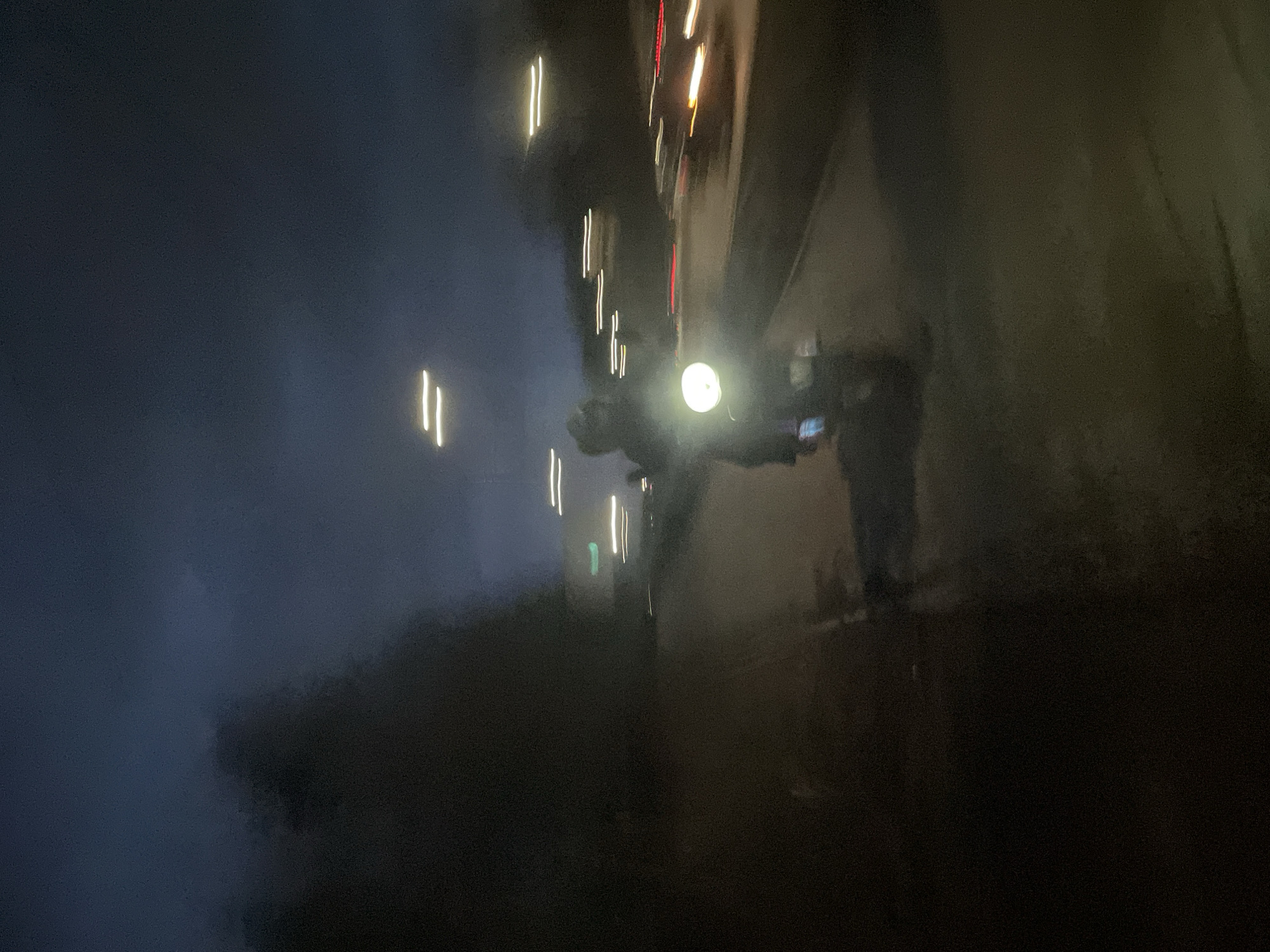 The image, likely taken at night and rotated, shows a dark cityscape with blurred light trails, possibly from vehicle traffic, suggesting motion blur due to camera movement or a long exposure setting. In the foreground, a person with a bright, circular light source is visible. They may be holding a phone or camera. Based on the context of the image and the mention of the city of Izhevsk, Russia, it is assumed that the location is within Izhevsk. The environment appears to be urban, characterized by buildings and streetlights. Further detail cannot be determined due to blur and low light.