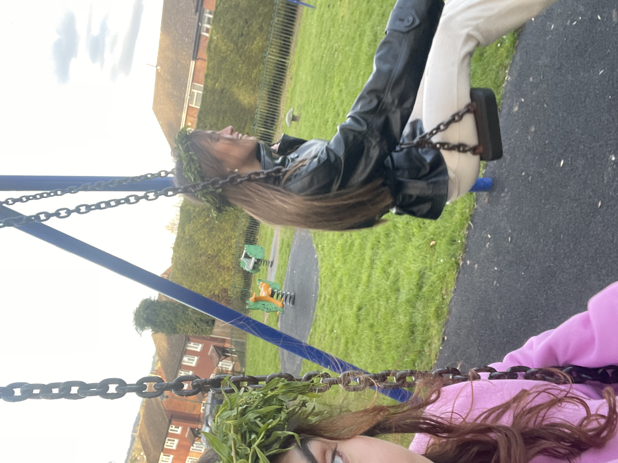 The image shows two individuals on swings in a playground setting. One person is wearing a black jacket and light-colored pants, positioned on a swing with a metal chain. The other is wearing a pink hoodie. Both are adorned with what appears to be greenery in their hair, possibly a wreath. In the background, there is a grassy area with spring riders, trees, and houses. The location is in Maidstone, United Kingdom.