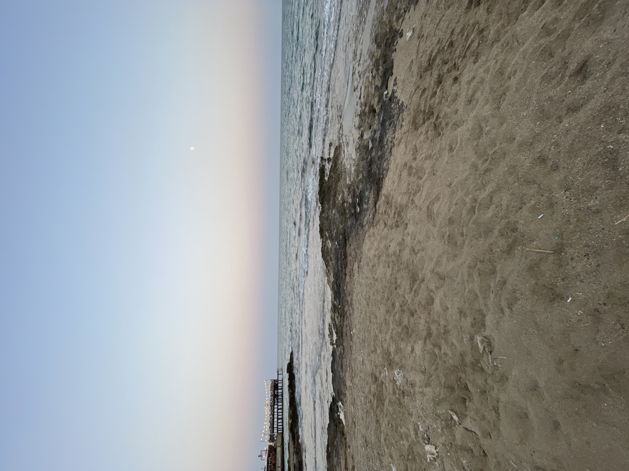 The image shows a beach scene at dusk in Trikomo, Northern Cyprus. The sand in the foreground is littered with debris, including plastic and small sticks. Rocky formations are visible along the shoreline, and the sea is calm with gentle waves. In the distance, a wooden pier extends into the water, illuminated by strings of lights, with a building at its end. The sky is a gradient of soft blues and pinks, with a faint moon visible in the upper right quadrant. The overall impression is one of a peaceful, yet somewhat neglected, coastal area as evening approaches.