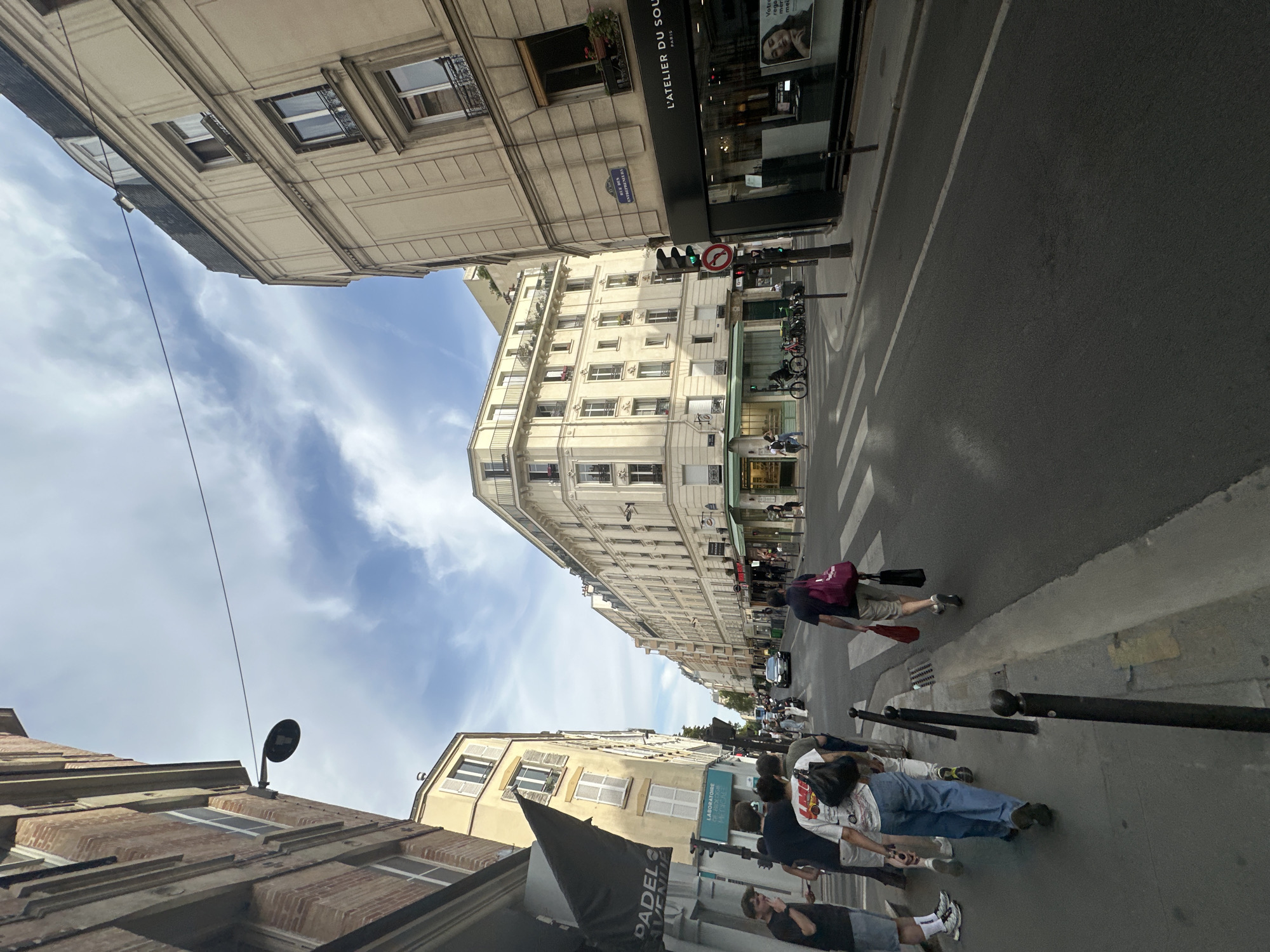 The image depicts a street scene in Paris, France. The perspective is from ground level, looking down a street lined with buildings on both sides. The sky is partly cloudy with blue patches.
On the left side of the street, there is a brick building with multiple windows and an awning. Further down, there are lighter-colored buildings with balconies and numerous windows. Several people are walking on the sidewalk on the left. One person is wearing a white t-shirt with a graphic print and blue jeans, carrying a black bag over their shoulder. Another person, a woman, is wearing a white t-shirt and blue jeans and is also carrying a bag. A person wearing a black t-shirt and denim shorts is standing to the far left.
On the right side of the street, there is a light-colored building with ornate architectural details, including moldings and balconies. A storefront with large glass windows displays products and a poster of a woman. The sign above the storefront reads "L'ATELIER DU SOUL... PARIS". A traffic light is visible, showing green, and a circular red and white sign indicating no entry. Further down the street, more buildings and shops are visible, with some pedestrians and parked bicycles. A car is driving down the street in the distance. The street has crosswalks and a sidewalk with black bollards. The overall impression is a busy urban street during the day.