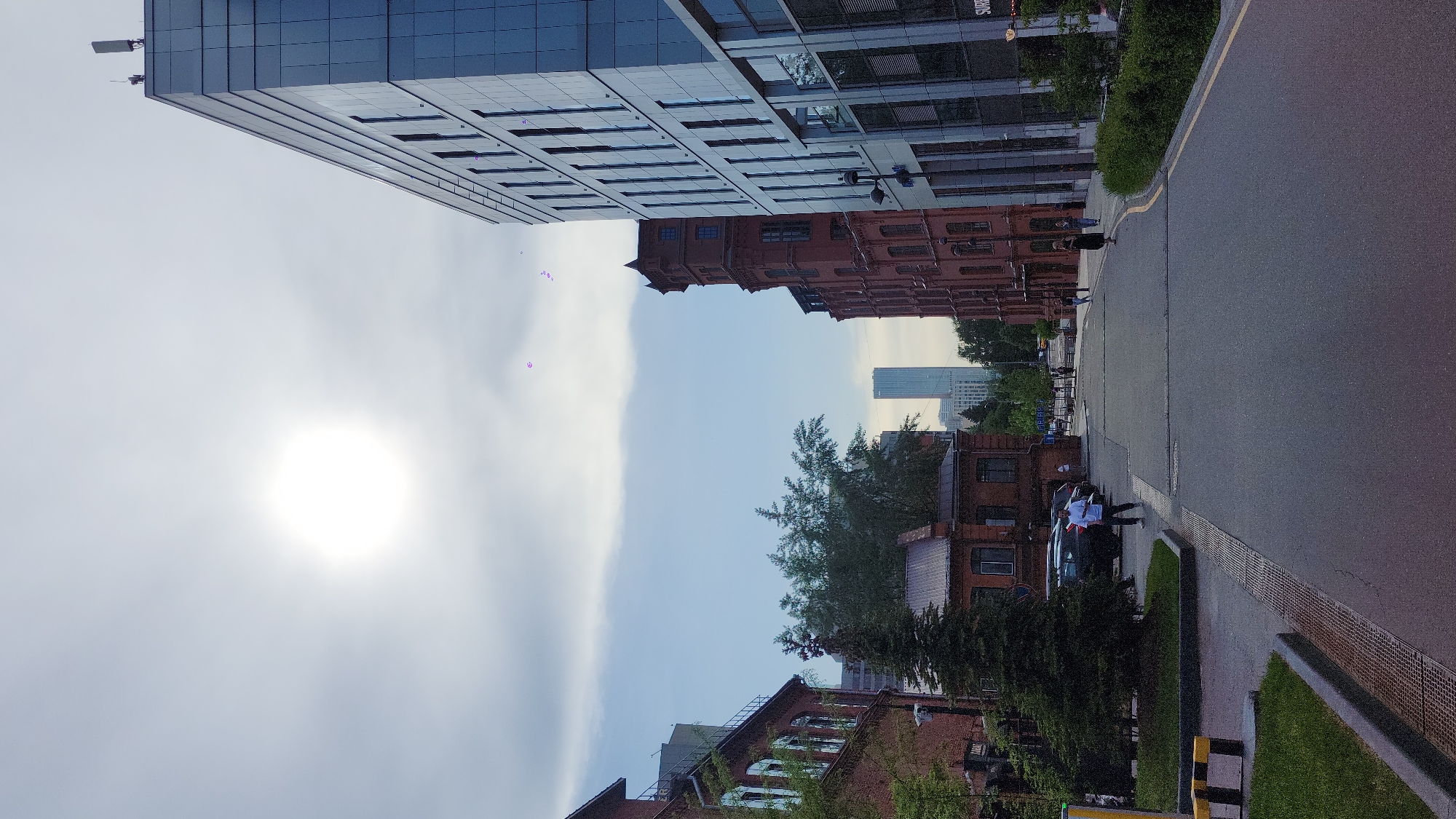 The image depicts a street scene in Moscow, Russia. The perspective is from the ground, looking upwards along a street lined with buildings of varying architectural styles. A modern glass-faced skyscraper dominates the upper-left portion of the image, while older red brick buildings are visible on the right side of the street. A tall, slender skyscraper is visible further down the street, between the buildings on either side. There are people walking along the pavement, and a parked car with people nearby. The sky is partly cloudy, with the sun obscured by the clouds.