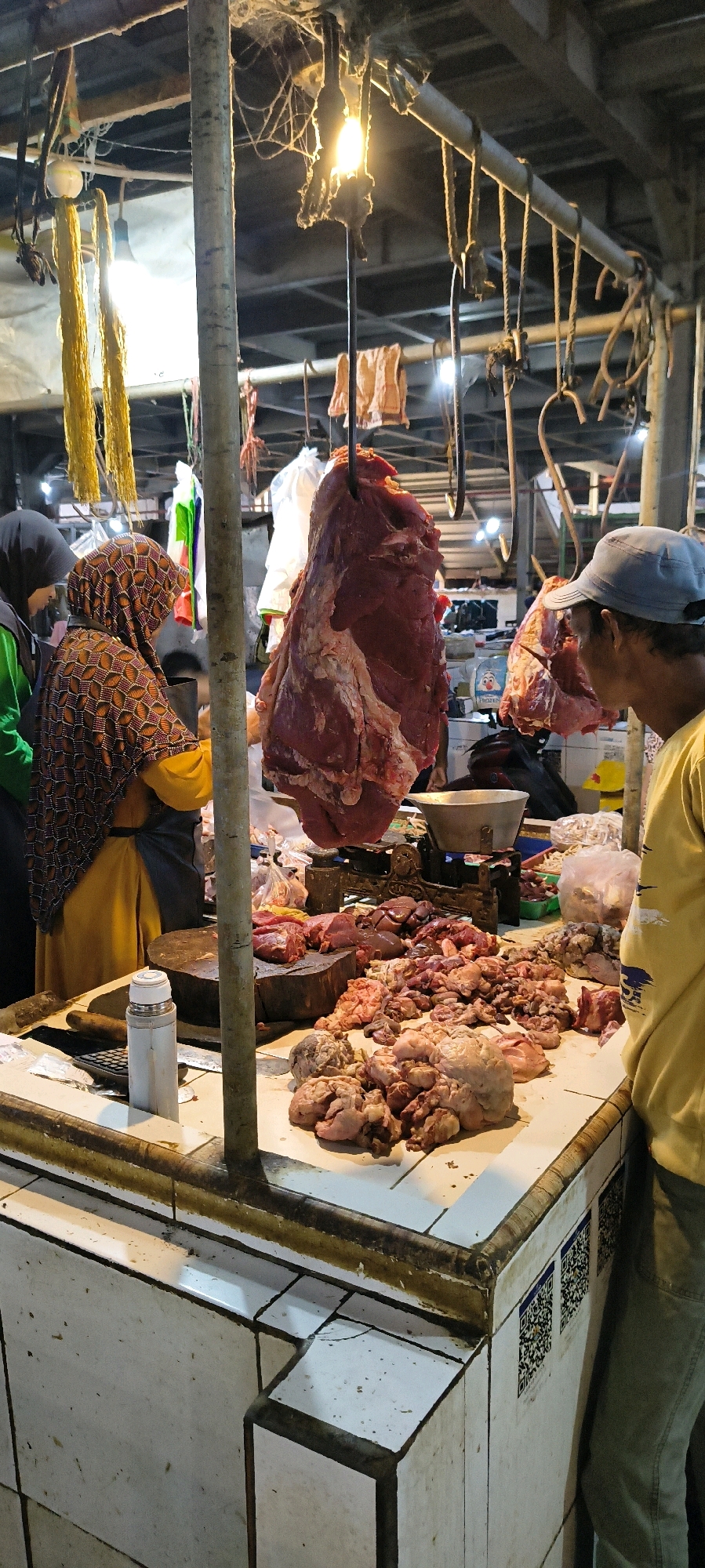 This image depicts a busy butcher stall in a market in Weru Lor, Indonesia. A large cut of raw red meat hangs from a hook above a counter laden with various smaller cuts of meat, offal, and other butcher items. Two women in headscarves, one in a yellow tunic and the other in a patterned headscarf and apron, are visible behind the counter, appearing to be serving or preparing meat. A man wearing a blue cap and a yellow shirt stands to the right, looking towards the meat. The stall is enclosed by a white tiled counter with a metallic trim and appears to be under a covered structure with visible lighting and hanging wires. The overall scene is one of commerce and food preparation in a local market setting.