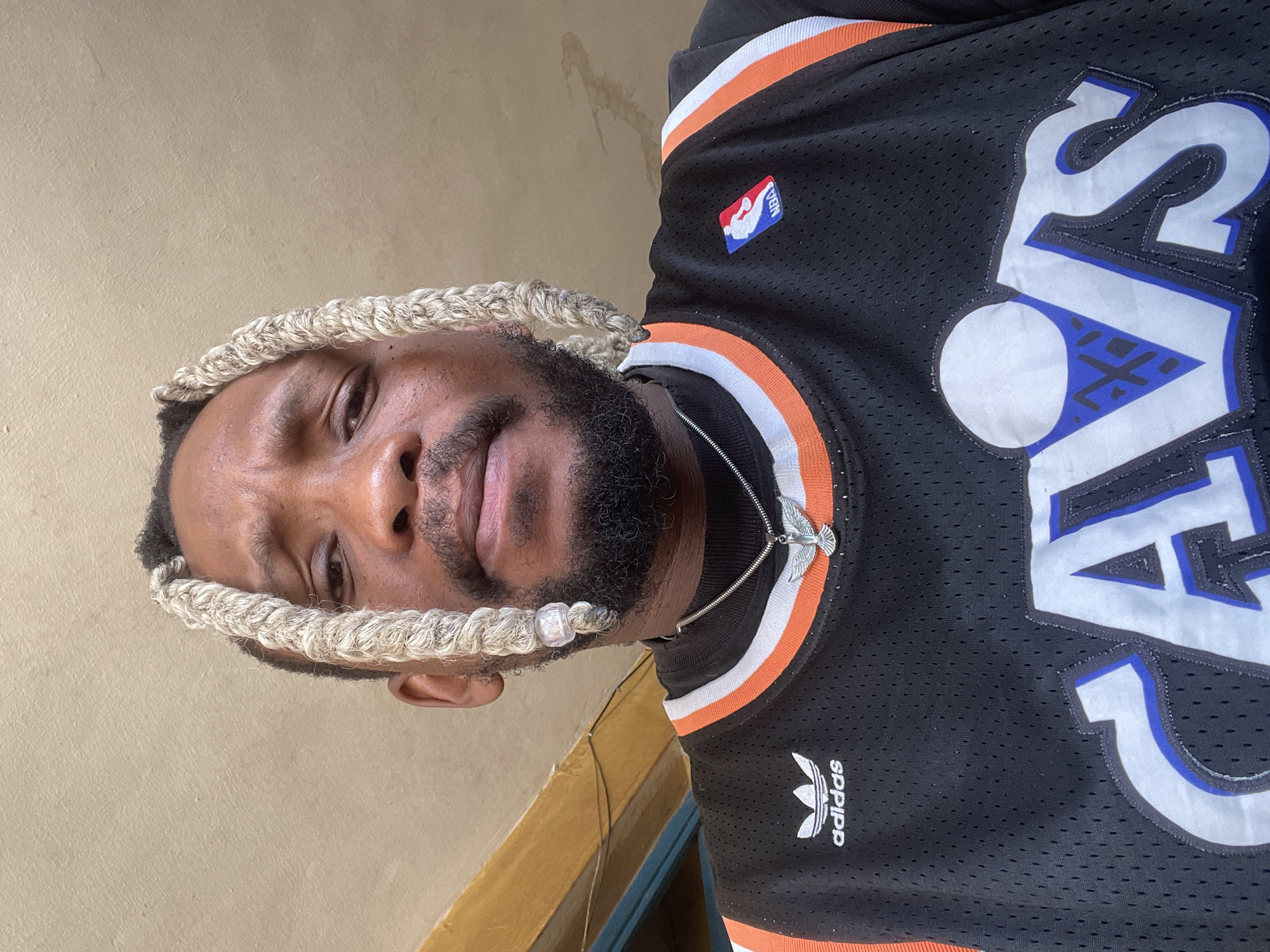 A portrait of a dark-skinned man wearing a black Cleveland Cavaliers basketball jersey, an Adidas logo is visible on the jersey. He has a full black beard and his hair is styled in blonde braids wrapped around his head and neck. He is wearing a silver chain. The picture was taken in Jos, Nigeria. The background is a beige colored wall, and the photo appears to be taken indoors.