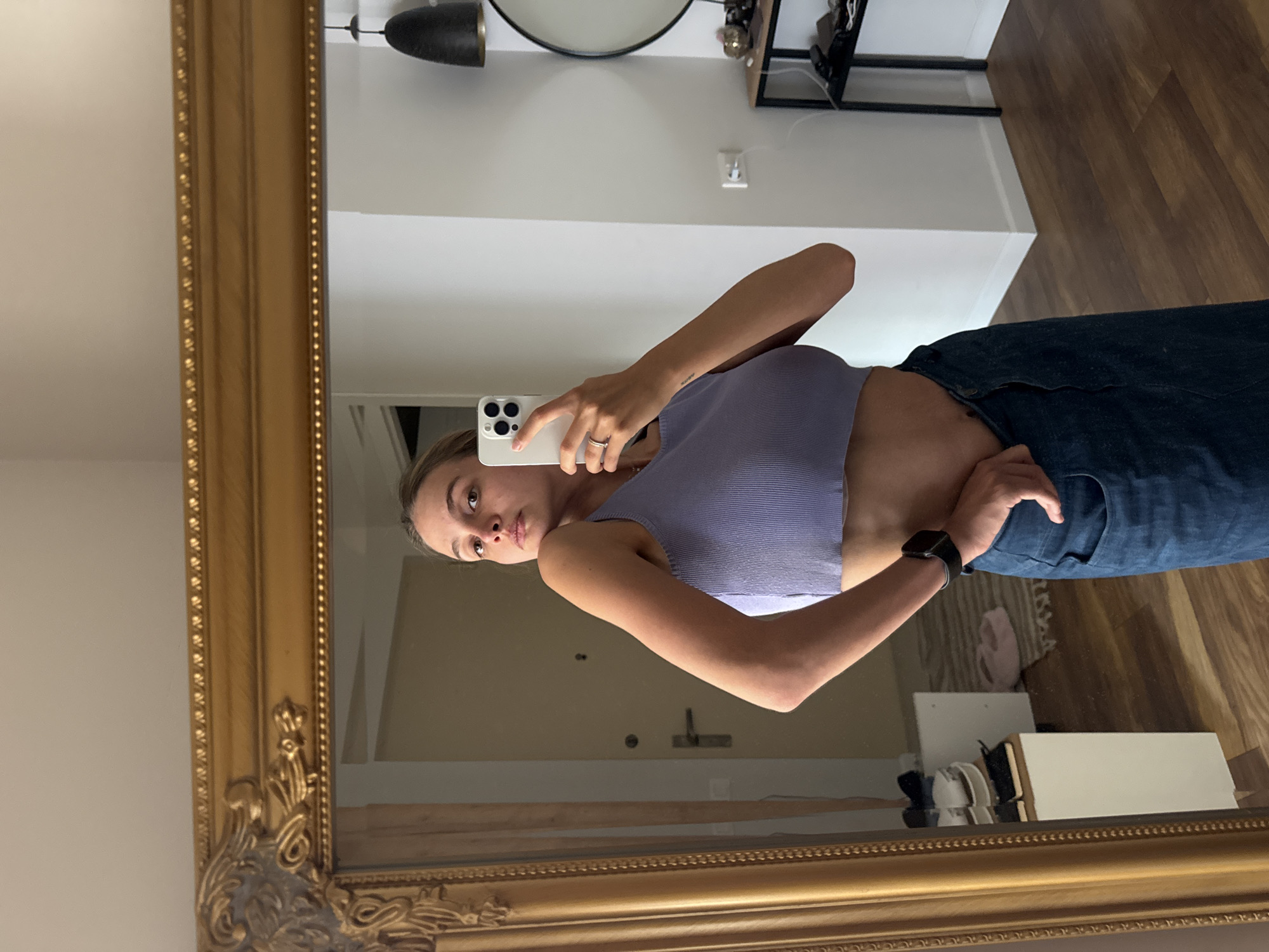 An image, originally rotated 90 degrees clockwise, captures a young woman taking a mirror selfie indoors. The woman, positioned centrally, is reflected in a large mirror with an ornate gold frame. She holds a white smartphone with multiple camera lenses in her left hand, which also bears a ring. Her right hand is casually tucked into the waist of her dark blue jeans. She is wearing a form-fitting, light purple ribbed crop top that exposes her midriff, and a dark-colored smartwatch is visible on her right wrist. Her expression is neutral, and she appears to be looking directly at the phone's camera.
The background visible in the mirror's reflection reveals elements of an interior space. A white wall occupies the upper portion, meeting a wooden floor. A black pendant light fixture and a round mirror are mounted on the wall above the woman's reflected head. Other reflected objects include a dark shelf unit with various items, and closer to the bottom, white cabinetry or furniture with some shoes or other objects on the floor next to it. The lighting is bright and even, suggesting an indoor setting like a bedroom or hallway. No discernible text is visible in the image.