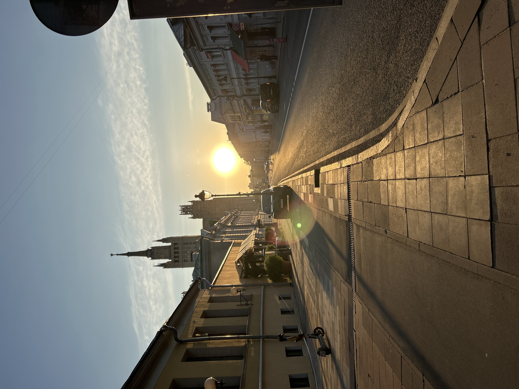 A ground-level, angled outdoor shot depicts a city street under a bright sun. The sky is partly cloudy, with a distinct contrail near the horizon. The road runs straight ahead, bathed in the sun's glare. Buildings line both sides of the street, featuring various architectural styles. A dark-colored car is visible on the road. On the right side of the frame, a paved sidewalk is present, with a parked scooter. The image is taken in Moscow, Russia.