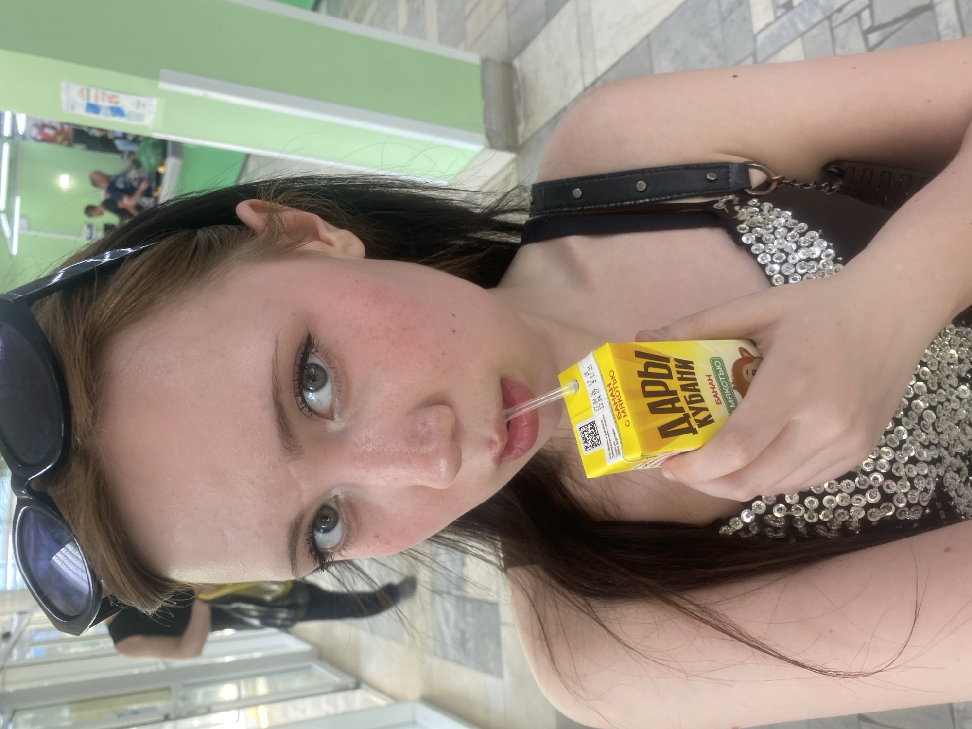 A young woman is drinking a yellow juice box with a straw. She is fair-skinned with long brown hair and is wearing sunglasses atop her head and a black top with silver sequins. The juice box is labeled in Cyrillic script. She appears to be indoors with tile flooring. In the background, people are visible through a window or opening. The location is Unknown, Unknown.