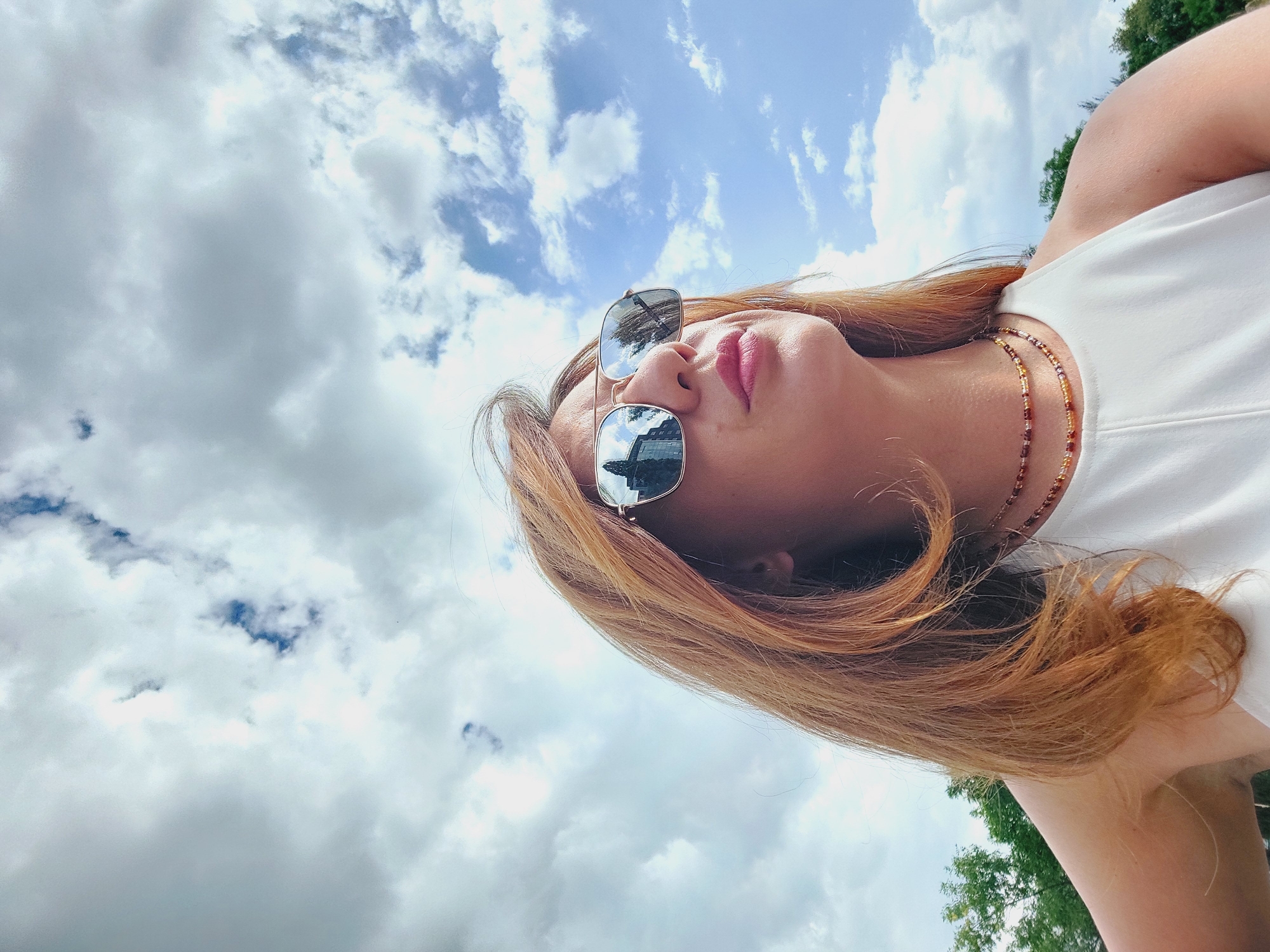 A medium shot photo shows a woman with auburn hair and sunglasses, looking up at a cloudy sky. She is wearing a white sleeveless top and two beaded necklaces. The sunglasses reflect buildings and trees. The scene appears to be outdoors, with greenery visible in the background. The location is the city of Sofia, Bulgaria.