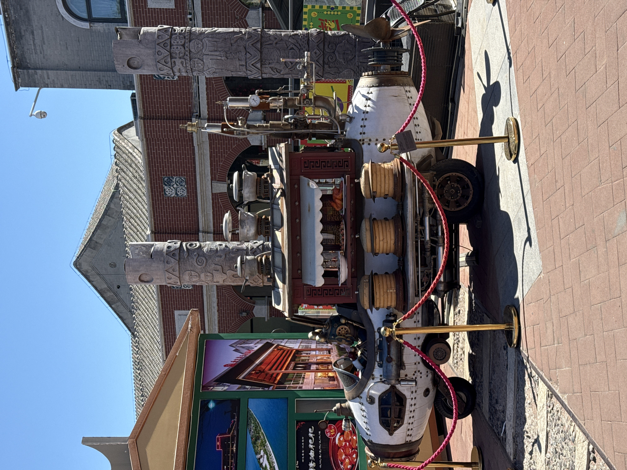 A street scene in Dongcheng District, China, featuring a complex, steampunk-style vehicle. The vehicle has a white front section and a variety of mechanical components, including gauges and pipes, situated on top. The street is paved with reddish-brown bricks, and the vehicle is roped off with red rope supported by golden poles. In the background, there are buildings constructed from red brick and gray stone, with architectural details that suggest traditional Chinese design elements, such as sloping roofs and carved wooden pillars. A large advertisement featuring images of local attractions is visible to the left of the vehicle. The sky is a clear blue. The shadow of a person holding up two fingers for a peace sign is cast on the ground.