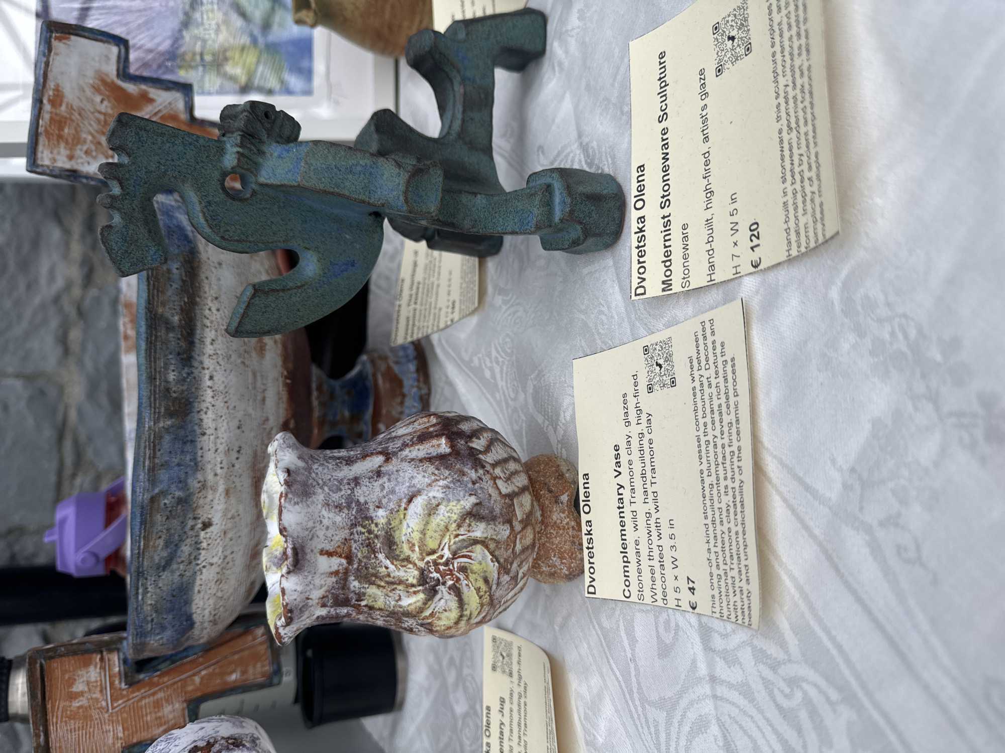This image depicts a display of handmade pottery and stoneware sculptures, likely at an art market or gallery in Kilkenny, Ireland. In the foreground, two cream-colored cards with black text describe the artworks. The left card identifies a "Complementary Vase" by Dvoretska Olena, made from stoneware with wild Tramore clay and glazes, wheel-thrown, hand-built, and high-fired. It measures H 5 x W 3.5 inches and is priced at €47. Beside it, a textured, earth-toned vase with swirling yellow and white glaze patterns is visible.
The right card describes a "Modernist Stoneware Sculpture" also by Dvoretska Olena. This piece is stoneware, hand-built, high-fired with an artist's glaze, measuring H 7 x W 5 inches and priced at €120. A teal-colored, abstract sculpture with interlocking shapes stands on a white cloth surface in front of this card.
In the background, larger ceramic pieces are partially visible, including a substantial blue and white patterned bowl. A purple plastic object rests on the rim of this bowl. The overall setting appears to be indoors, possibly with natural light filtering in, given the bright areas and shadows. The display suggests a curated presentation of artistic ceramics.