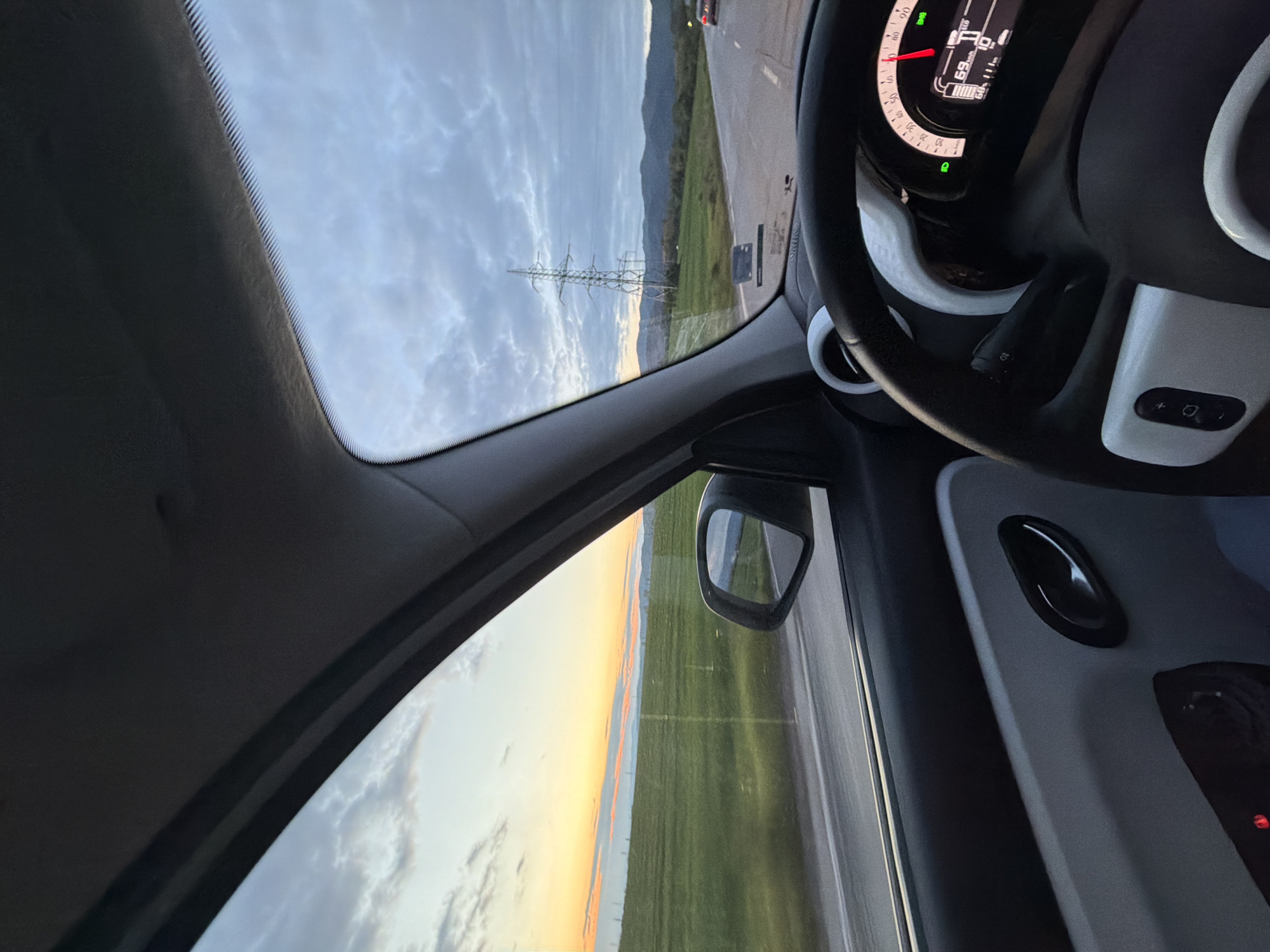 The image shows the interior of a car, specifically the driver's side, with a view through the windshield and side window. The dashboard and steering wheel are visible, with a speedometer reading 69 km/h. Outside the windshield, there's a road, grassy fields, mountains, and power lines under a cloudy sky. The side window shows a road and a green field, likely during sunset or sunrise due to the orange glow on the horizon. The car is presumably driving on a road within or near the city of Buhovo, Bulgaria, given the location.