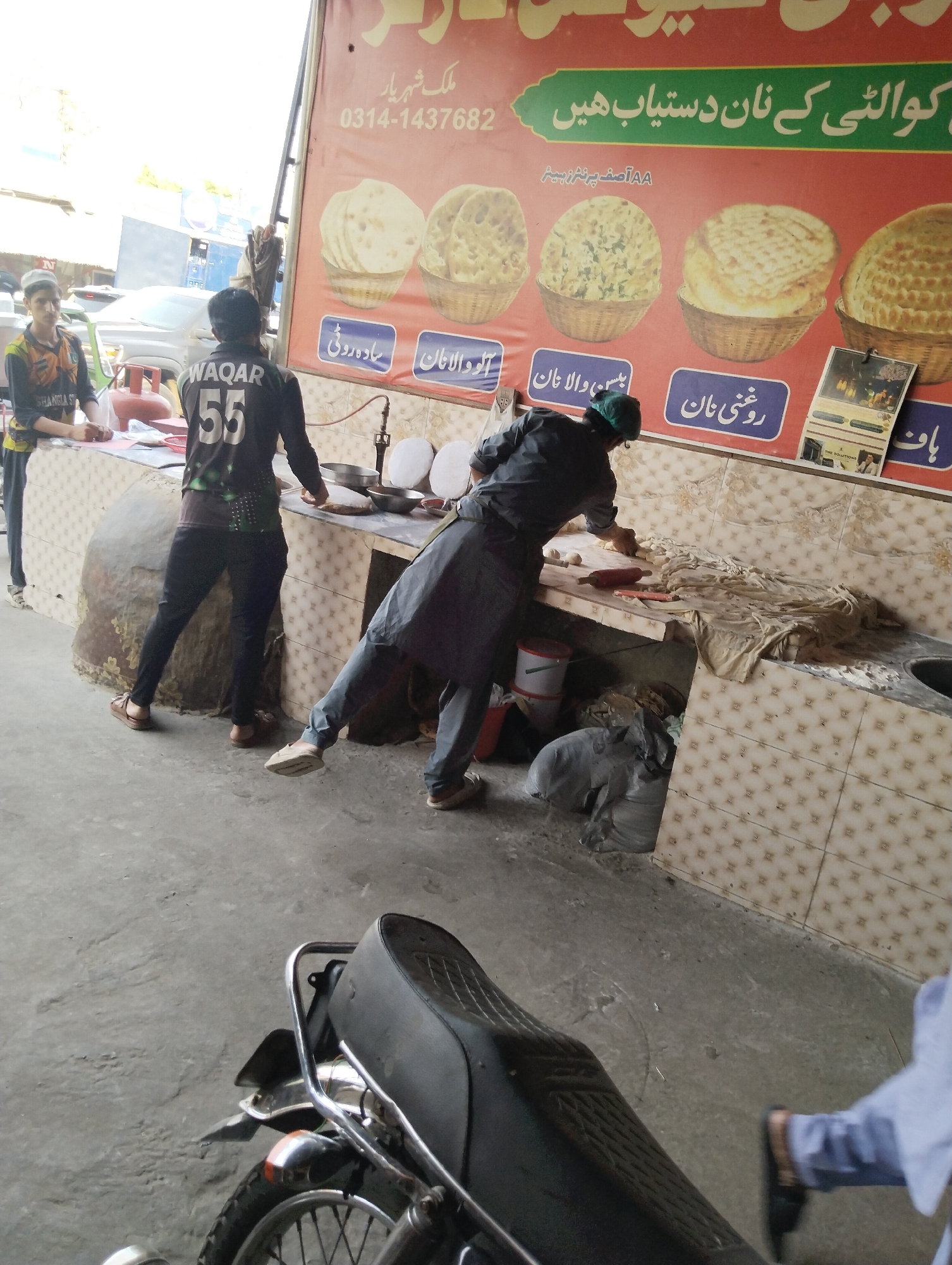 The image depicts a bustling street-side food stall, likely a nanbai (bread maker's) shop, captured during daylight hours in Heir, Pakistan. The scene centers on the preparation of various types of bread, with three men actively involved or present.
In the foreground, the black seat and chrome handlebars of a motorcycle are partially visible, suggesting a busy street environment. The background shows blurred outlines of vehicles, including a white car and a blue truck, indicating a roadside location.
On the left, a young man wearing a white cap and a green and black long-sleeved shirt with "SHANGLA ST" printed on it, stands behind a counter. In the center, another man, sporting a black long-sleeved shirt with "WAQAR 55" on its back, leans over a tiled counter, engaged in dough preparation. To his right, a third man, wearing a dark outfit and a green cap, is actively kneading or shaping dough on a similar flour-dusted counter, with a rolling pin nearby. A large, stone tandoor-style oven is partially visible on the right, indicating traditional baking methods.
A prominent red and green banner above the counter advertises the shop. The Urdu text on the banner reads "کوالٹی کے نان دستیاب ھیں" (Quality naan available here) and "ملک شہر یار" (Malik Shahryar), along with a phone number "0314-1437682" and printer credit "AA آصف پرنٹر زبیر". The banner also displays images of different bread types, labeled "سادہ روٹی" (Plain Roti), "آلو والا نان" (Potato Naan), "بس والا نان" (Gram Flour Naan), and "روغنی نان" (Oily Naan), with a partially visible fifth option labeled "پاف" (possibly "Peshawari Naan"). A small calendar or poster that says "THE SOLUTIONS" hangs near the right side of the banner.
The work surfaces are covered with flour and dough, and various metal bowls, buckets, and a red gas cylinder are visible, highlighting the functional nature of the bread-making operation. The overall ambiance is that of a diligent and active street food vendor.