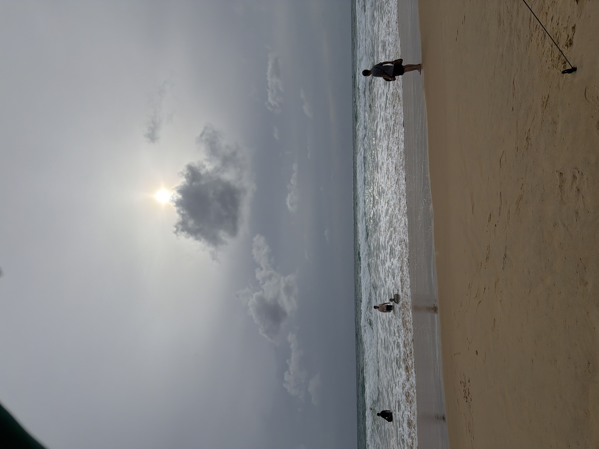 In Karon, Thailand, a beach scene unfolds under an overcast sky with a hint of sun breaking through the clouds. The foreground is dominated by tan sand, marked with faint footprints. The middle ground features the ocean with moderate waves, where several individuals are present.
On the left, near the shore, a person is sitting in the water, possibly with a surfboard. Further out in the water, another individual stands waist-deep. To the right, a person stands on the sandy shoreline, facing away from the camera, carrying a bag over their shoulder. A dark thin rope is visible in the lower right corner of the frame, anchored to a small black object. The overall mood is serene, with the natural elements of sand, sea, and sky taking center stage.