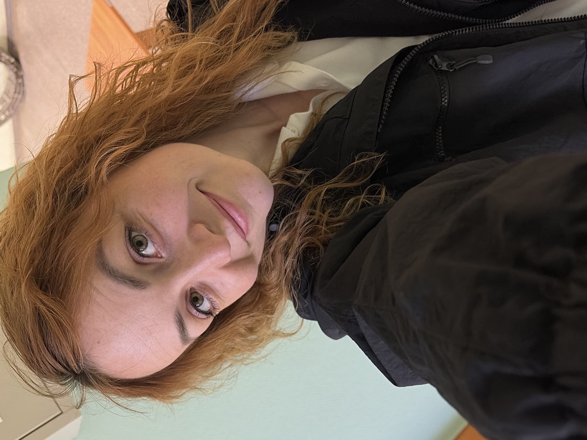 The image is an upward-angled selfie of a woman with long, wavy, reddish-brown hair, presumably taken in Chaikovsky, Russia. Her head is tilted to the side, and she looks directly at the camera with a subtle, friendly smile. Her eyes are light-colored, possibly green or hazel, with visible eyeliner and mascara.
She is wearing a light-colored, likely white, collared shirt or blouse underneath a dark, possibly black, jacket. The jacket features visible zippers, suggesting a casual or outdoor style.
The background indicates an indoor setting. A light, pastel green wall occupies a significant portion of the frame, particularly towards the bottom and right. In the upper left, a light brown wooden surface, possibly a piece of furniture, is visible. A blurry, metallic, and somewhat ornate object, perhaps a decorative item or part of a fixture, is faintly discernible on the far left. The lighting is soft and even, consistent with indoor conditions. There is no visible text in the image.