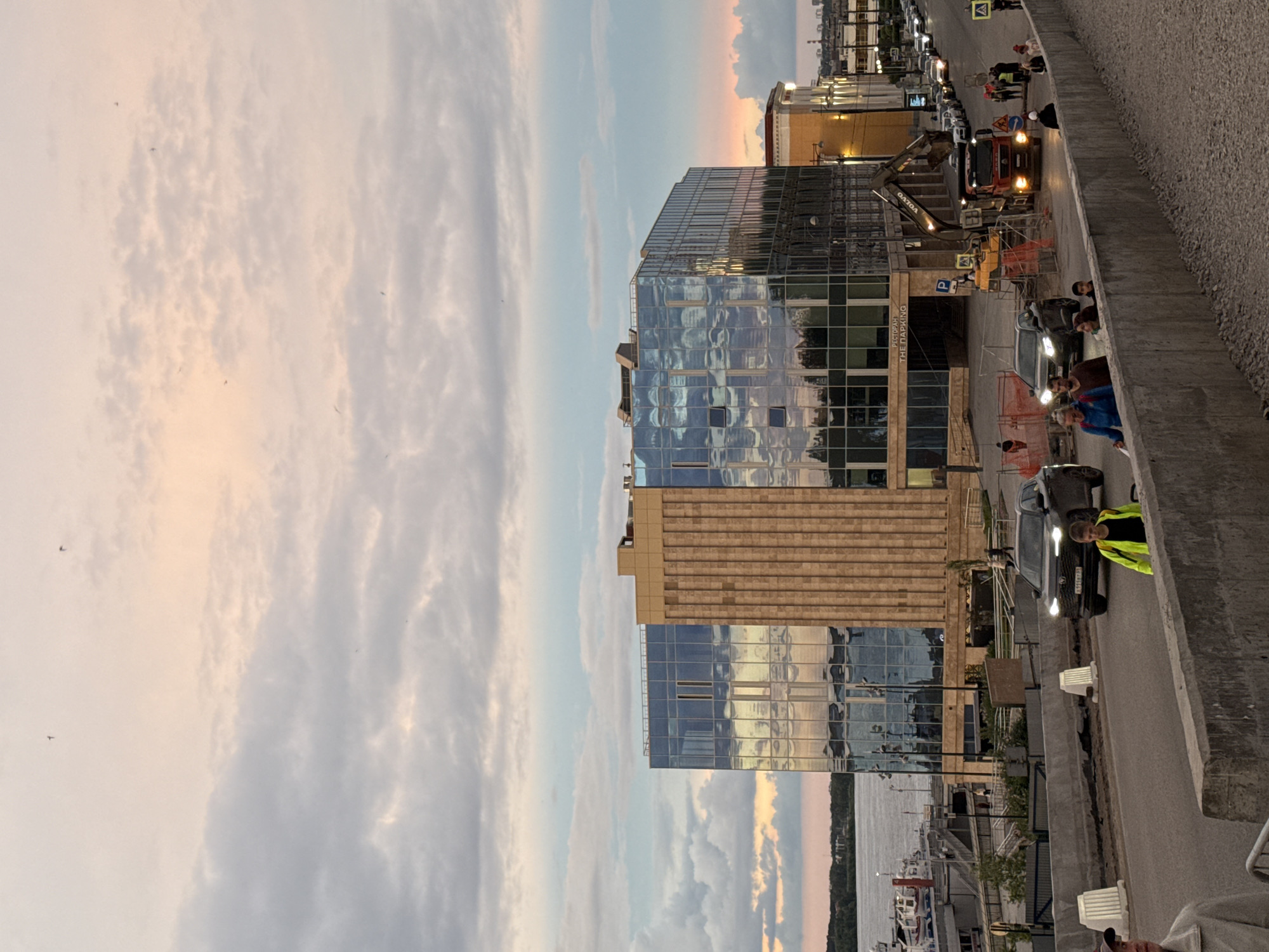 The image depicts a street scene in Perm, Russia, at dusk. A modern glass and tan stone building reflects the sunset sky. A gray concrete barrier separates the street from the sidewalk, where several people are standing and looking towards a work zone. In the work zone, there is an excavator, a red truck, and orange safety barriers. The road has parked cars on one side and active traffic on the other. A body of water is visible in the distance, reflecting the sunset colors. The sky is filled with soft clouds.