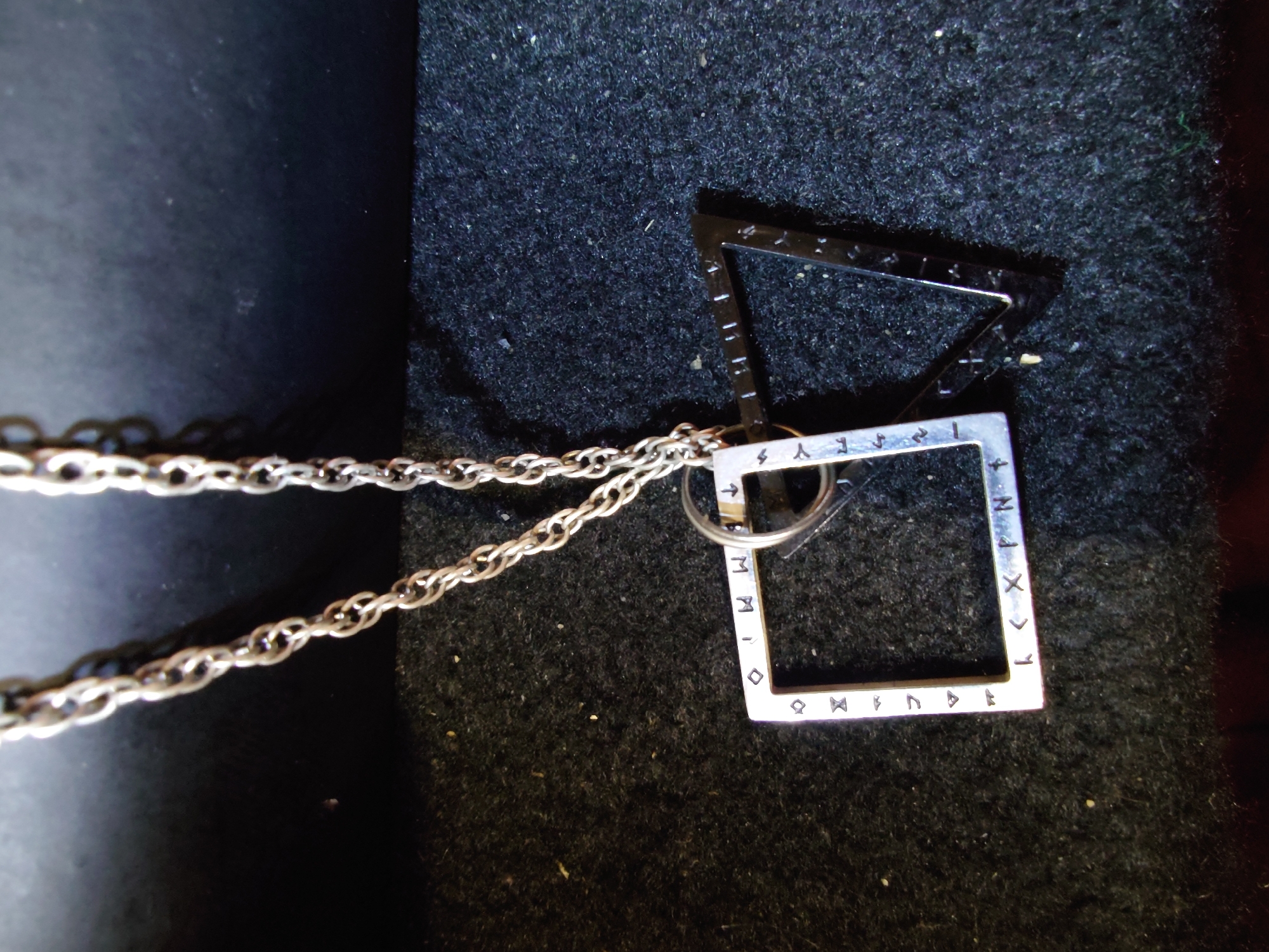 A close-up, eye-level shot captures a silver necklace with two pendants against a textured dark surface. The necklace has a fine, twisted chain. The left pendant is a silver square outline, with a darker silver inner border, inscribed with a series of small symbols that appear to be runes along its edges. The right pendant is a dark, possibly black metal, triangular outline. The overall lighting is dim, with a slight highlight on the chain and the silver pendant. The background consists of a dark, fuzzy material, likely fabric or felt, and a smoother, darker surface above. There are no people or animals visible. There is no indication of the city of Sokolohirsk, Ukraine in the image.