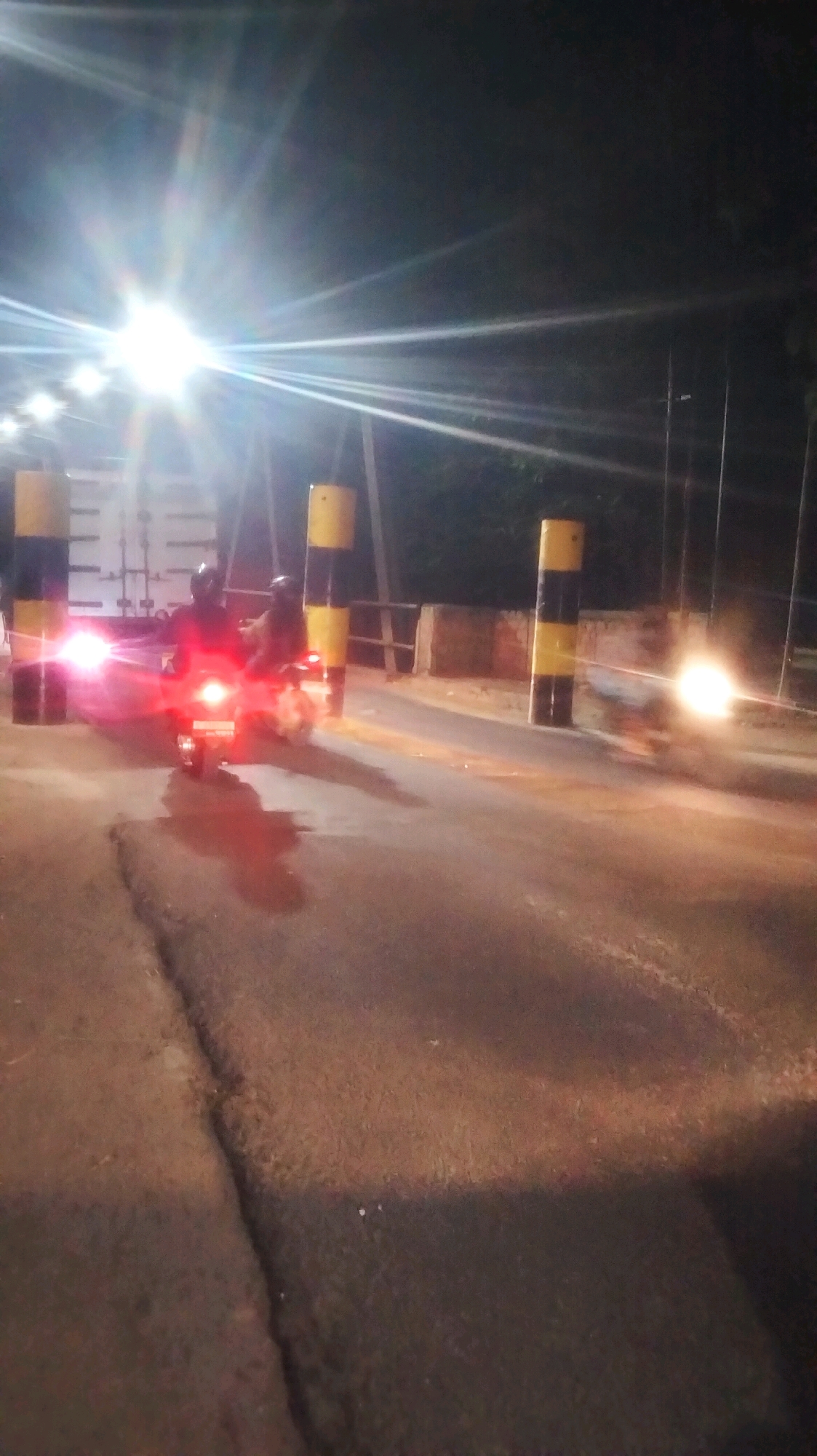 The image depicts a night scene on a road, possibly a toll gate or checkpoint, in Bojonegoro, Indonesia. Two individuals are riding a motorcycle, illuminated by the motorcycle's headlight and taillight, which cast a red glow on the road. The road surface appears to be cracked and worn. Yellow and black striped posts are visible on either side of the road, suggesting a barrier or lane divider. Bright lights, possibly streetlights or overhead illumination, create lens flares and streaks across the dark sky. In the background, trees and foliage are dimly visible. The overall impression is one of a typical night scene on a functional road in the mentioned location.