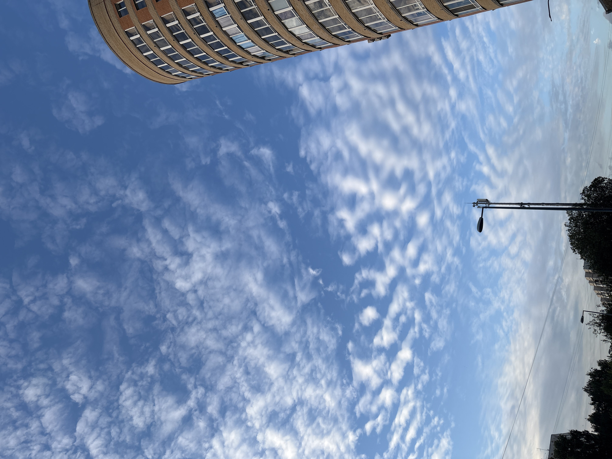 The image captures a view of a circular brick building with numerous windows against a bright blue sky filled with wispy white clouds. A tall, dark lamppost with a communication antenna on top stands in the foreground. Below the sky, a line of trees and other buildings are visible at the bottom of the frame, suggesting an urban setting. The scene is likely from Saint Petersburg, Russia, given the architectural style of the building and the characteristic cloud formations often seen in the region. The image appears to be taken during daylight hours, possibly late afternoon or early morning, given the soft lighting. There are no people or animals visible in the image. The overall impression is one of a serene urban landscape.