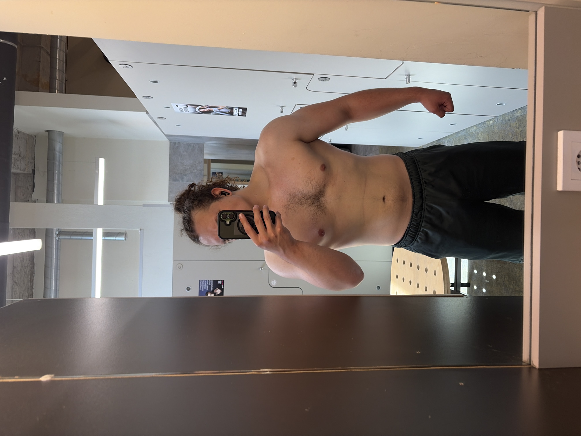 A shirtless man is taking a selfie in a locker room. The man is wearing dark-colored athletic pants. The background shows lockers and a neutral-toned interior. The setting appears to be a gym or fitness facility. The specific location in Plovdiv, Bulgaria, cannot be determined from the image.
