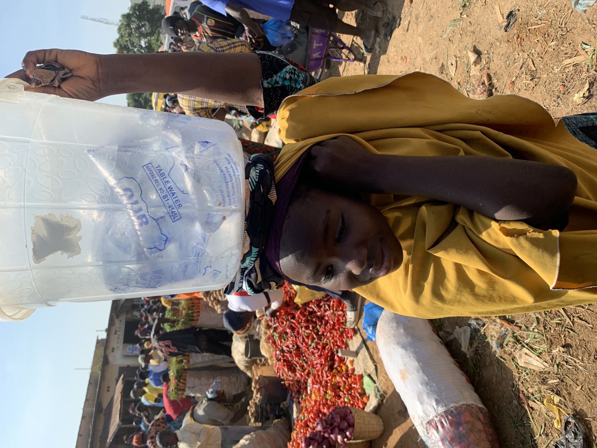 In the city of Gura Top, Nigeria, a young girl with dark skin and wearing a yellow headscarf and robe balances a clear plastic bucket filled with water on her head. She is in a bustling outdoor market. Her right arm is raised, supporting the bucket. The bucket has blue lettering and markings on it. Around her, the market is crowded with people. In the foreground, there are large piles of red chili peppers and some red onions in baskets. Further back, there are sacks of produce and more market stalls with people browsing and selling goods. The ground is dusty and dry, typical of an outdoor market environment. The sky is clear and bright, indicating a sunny day.