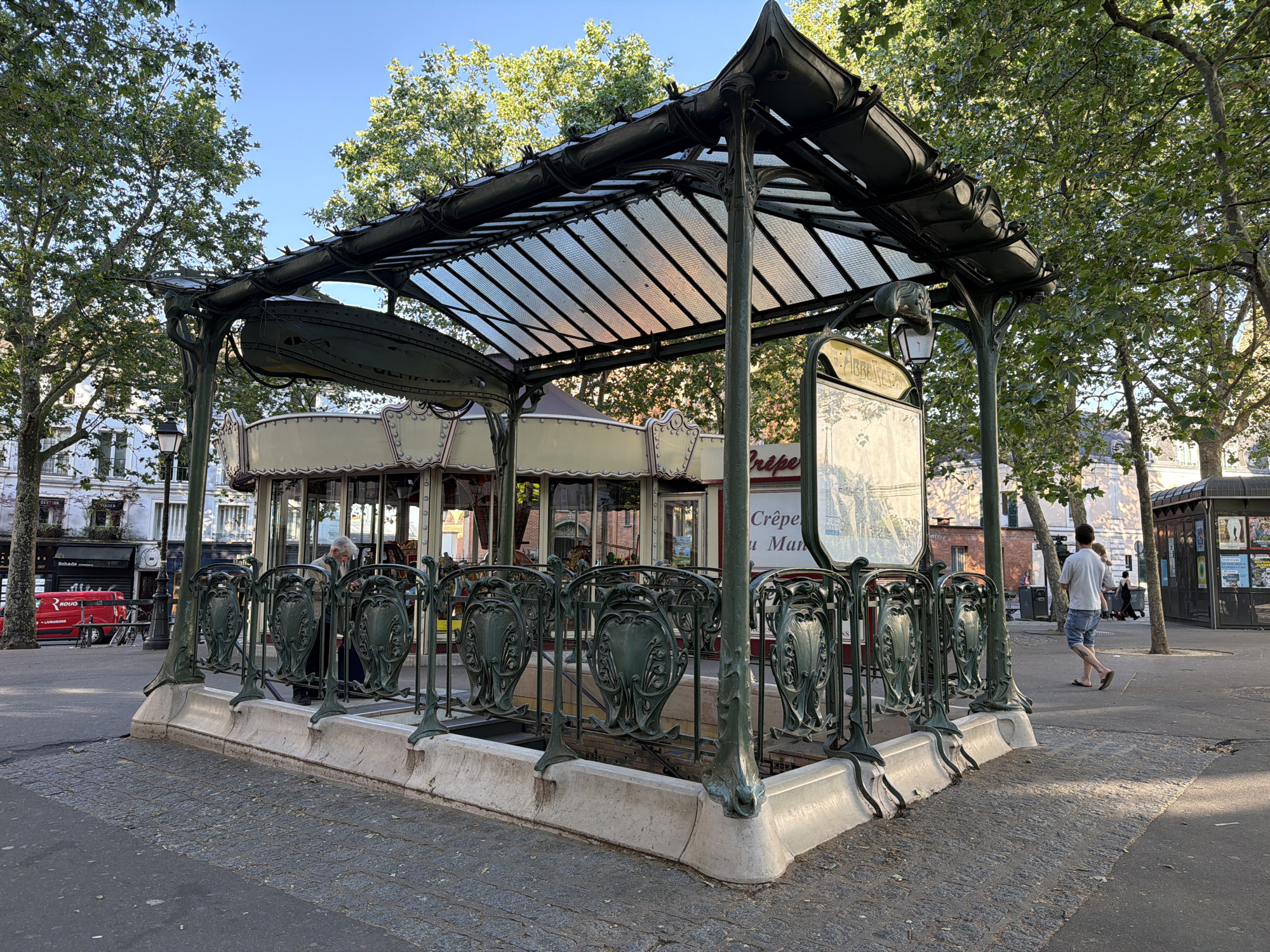 The image captures a classic Parisian metro entrance with Art Nouveau details. The entrance is enclosed by a decorative metal railing and sheltered by a glass roof supported by ornate metalwork. In the background, a carousel adds a whimsical touch to the scene. People are walking by and the surrounding area includes trees, buildings, and a newsstand, typical elements of a Parisian streetscape. Based on the visible signage, the location is near the Abbesses station in Montmartre, Paris, France.