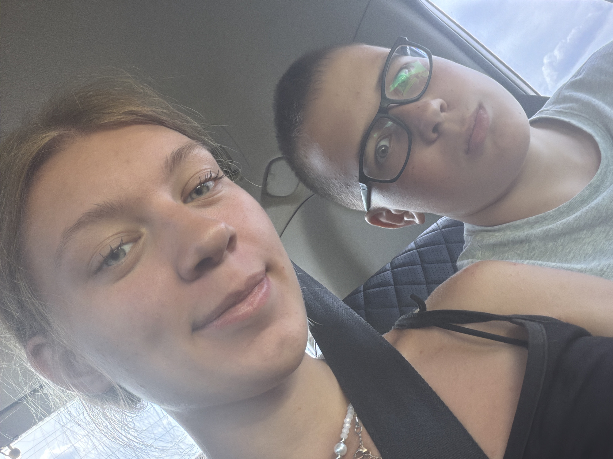 Two individuals, a young female and a young male, are in the front seats of a vehicle, likely a car, as indicated by the presence of seatbelts and car interior elements. The female is in the foreground, angled towards the camera, with her blonde hair visible. She is wearing a black top and a necklace with white beads. The male is positioned slightly behind and to the right of the female. He is wearing glasses with black frames and a grey t-shirt. The background shows the interior of the vehicle, including a quilted seat, and a glimpse of the outdoors through a window, suggesting daylight. The image is a selfie taken from a low angle. The location is identified as Novoe Feizullovo, Russia, though no specific landmarks or identifying features of this location are visible within the image itself.