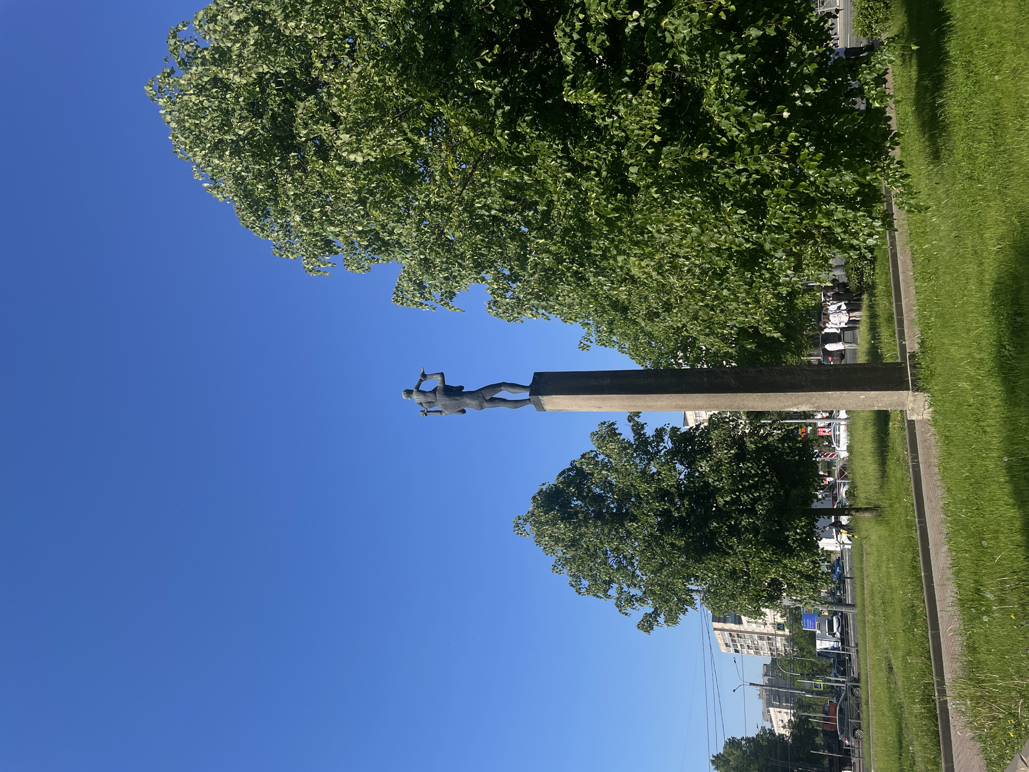 The original image is presented rotated 90 degrees clockwise. Describing it in its correct vertical orientation, the photograph captures a bright, sunny outdoor scene, likely a city park or boulevard alongside a street.
A vast, clear blue sky fills the upper half of the frame, suggesting midday or early afternoon conditions with bright sunlight. The central focus is a tall, dark-colored monument. It comprises a substantial rectangular pedestal topped with a human figure, sculpted from what appears to be bronze or dark metal. The figure is a stylized male athlete depicted in a dynamic pose, holding two disc-like objects or weights, resembling a discus thrower or shot putter.
Lush green trees with dense foliage are prominent, with one large tree framing the upper left portion of the sky and another appearing behind and to the right of the monument. To the right of the monument, a wide strip of manicured green grass runs along a grey paved curb or path.
In the background, on the far left side, an urban environment is visible. Several multi-story buildings line a street busy with cars and other vehicles. Utility poles and power lines are faintly discernible. There is no discernible activity beyond the static scene. No readable text is visible on the monument or elsewhere. The image portrays a public space where nature and urban elements converge around a commemorative statue.